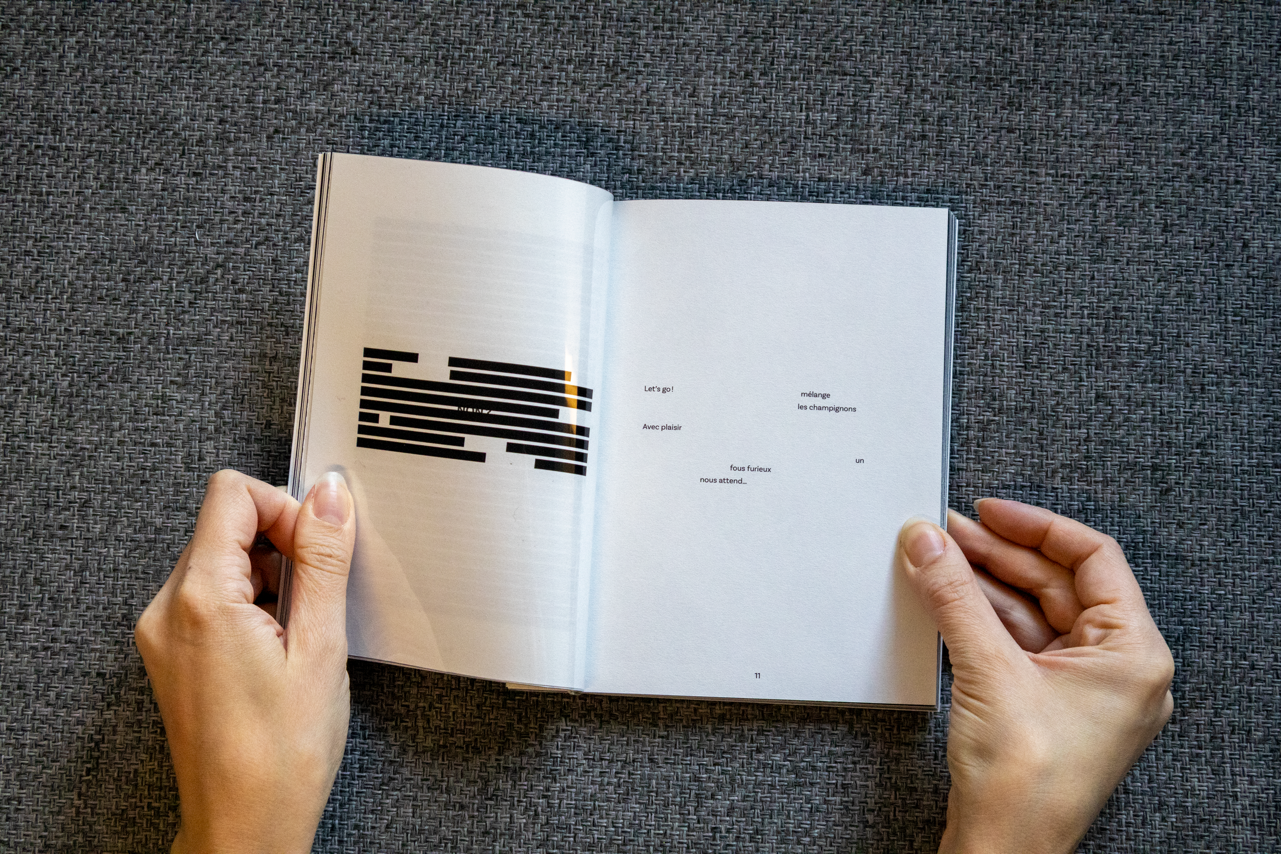 Une personne tient un livre ouvert avec des pages blanches et du texte en français, reposant sur une surface en tissu gris. La page gauche comporte une illustration graphique abstraite, la droite contient un poème ou texte en français.
