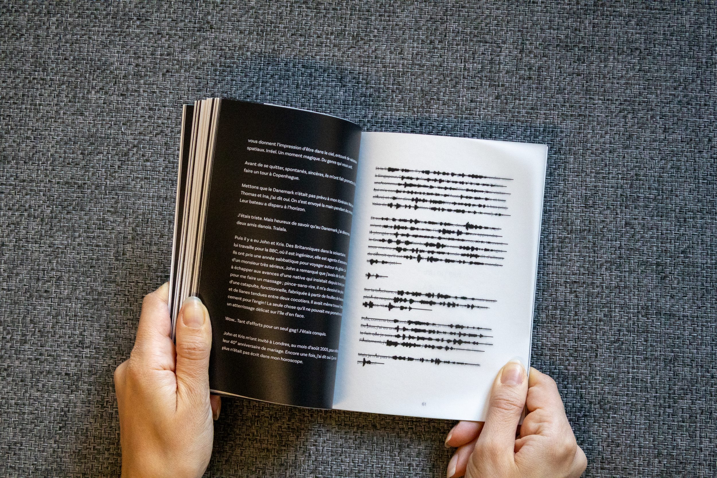 Une personne tient un livre ouvert avec du texte en français sur la page de gauche et une représentation graphique de lignes de temps ou de sons en noir sur la page de droite, posé sur une surface en tissu gris.