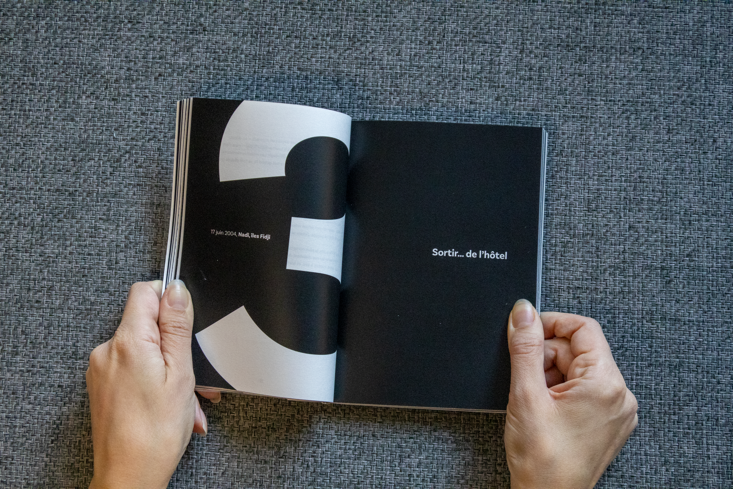 Une personne tient un livre ouvert avec une page noire et un texte blanc, sur une surface gris clair. La page gauche affiche un texte en français, la page droite a une phrase en français. Il s'agit d'un livre avec une conception moderne et minimalist