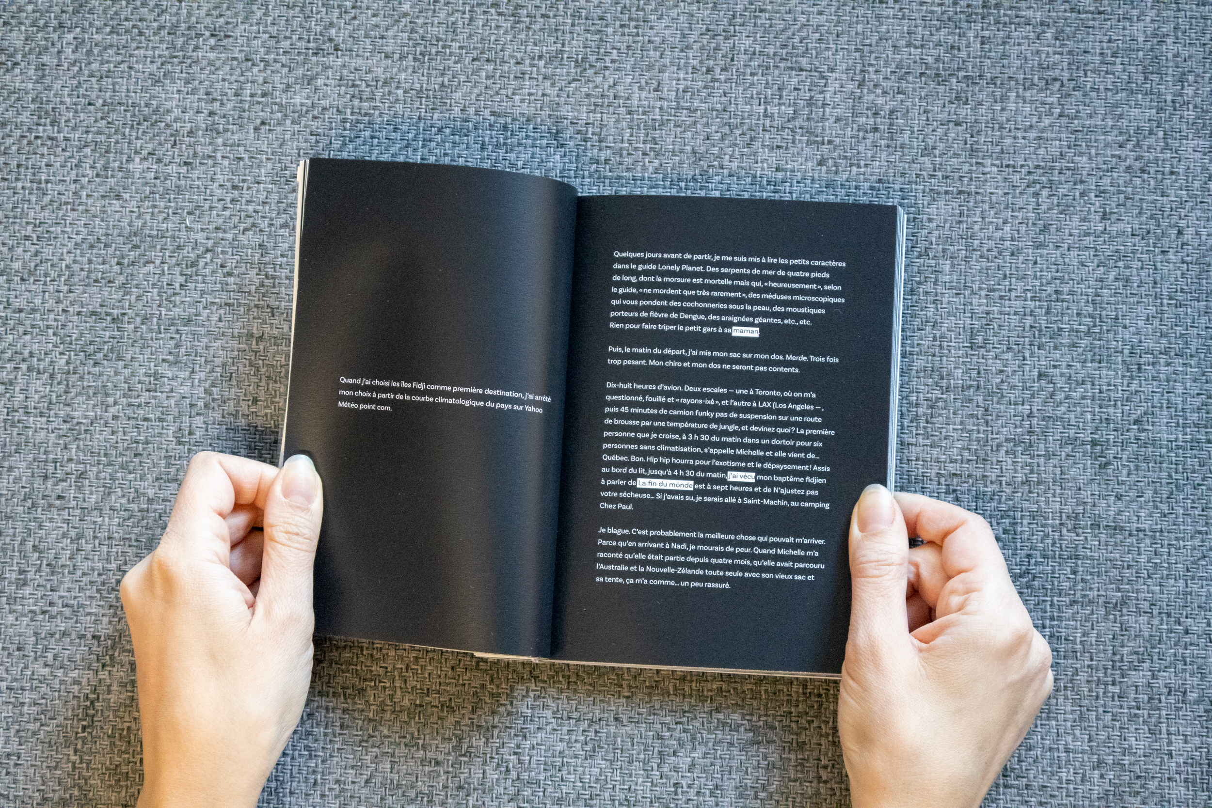 Une personne tient un livre ouvert avec des pages noires et du texte blanc, posé sur une surface recouverte d'un tissu gris