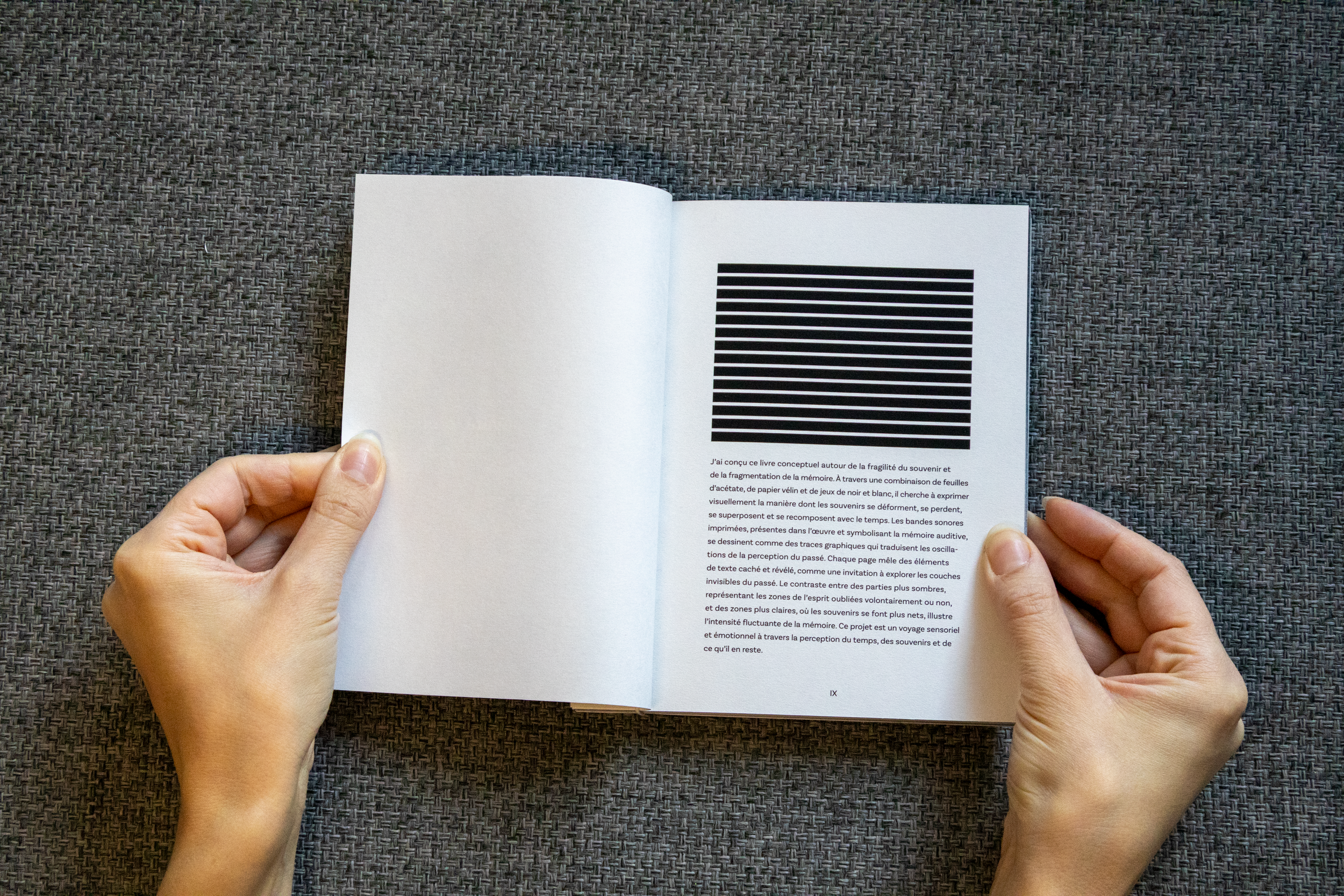 Une personne tenant un livre ouvert avec une page contenant une illustration composée de lignes horizontales noir et blanc et du texte en français en dessous, posé sur une surface en tissu gris.