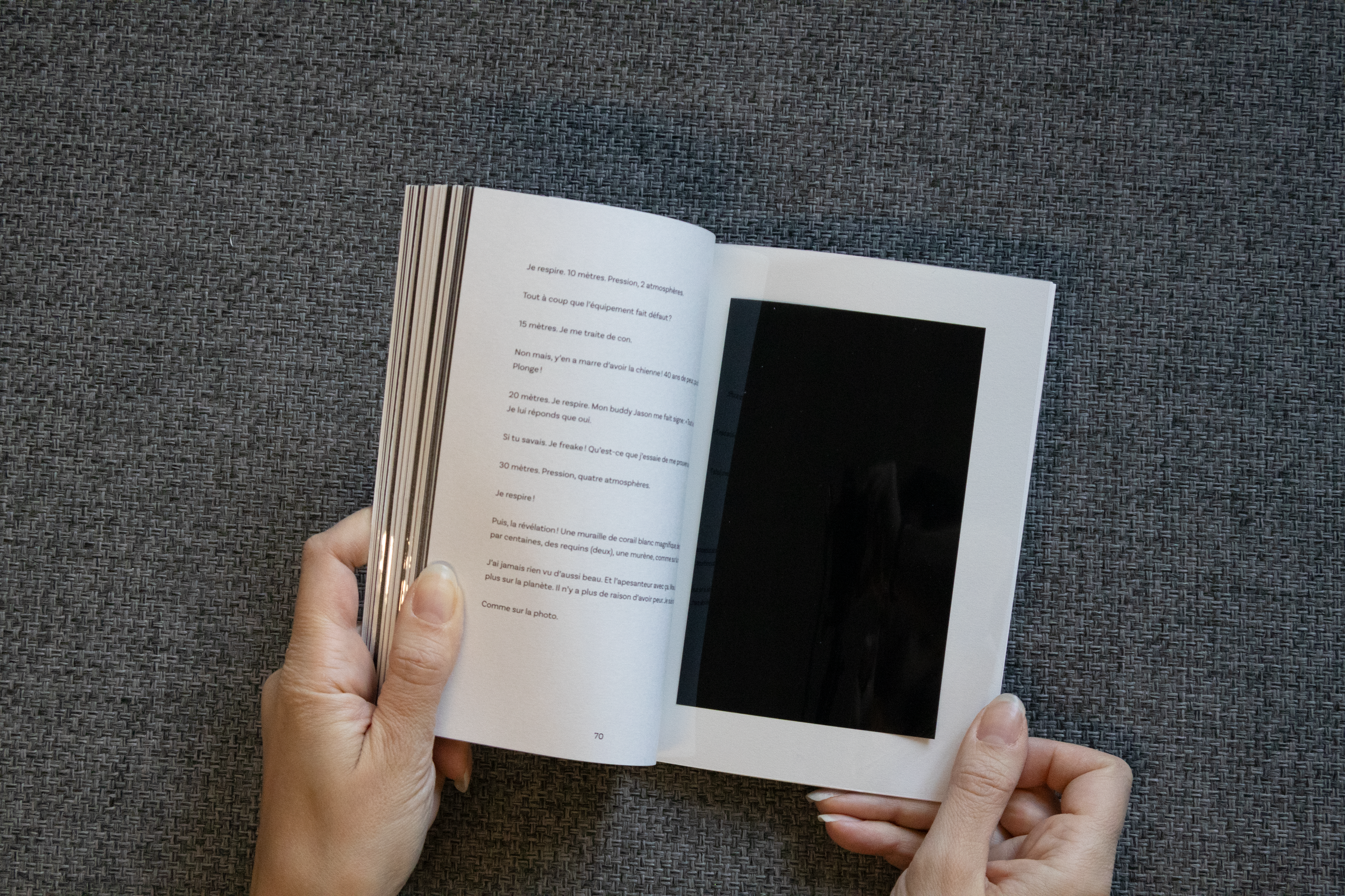 Une personne tient un livre ouvert avec du texte en français sur la page de gauche et une grande image noire sur la page de droite, le tout posé sur un fond gris en tissu.