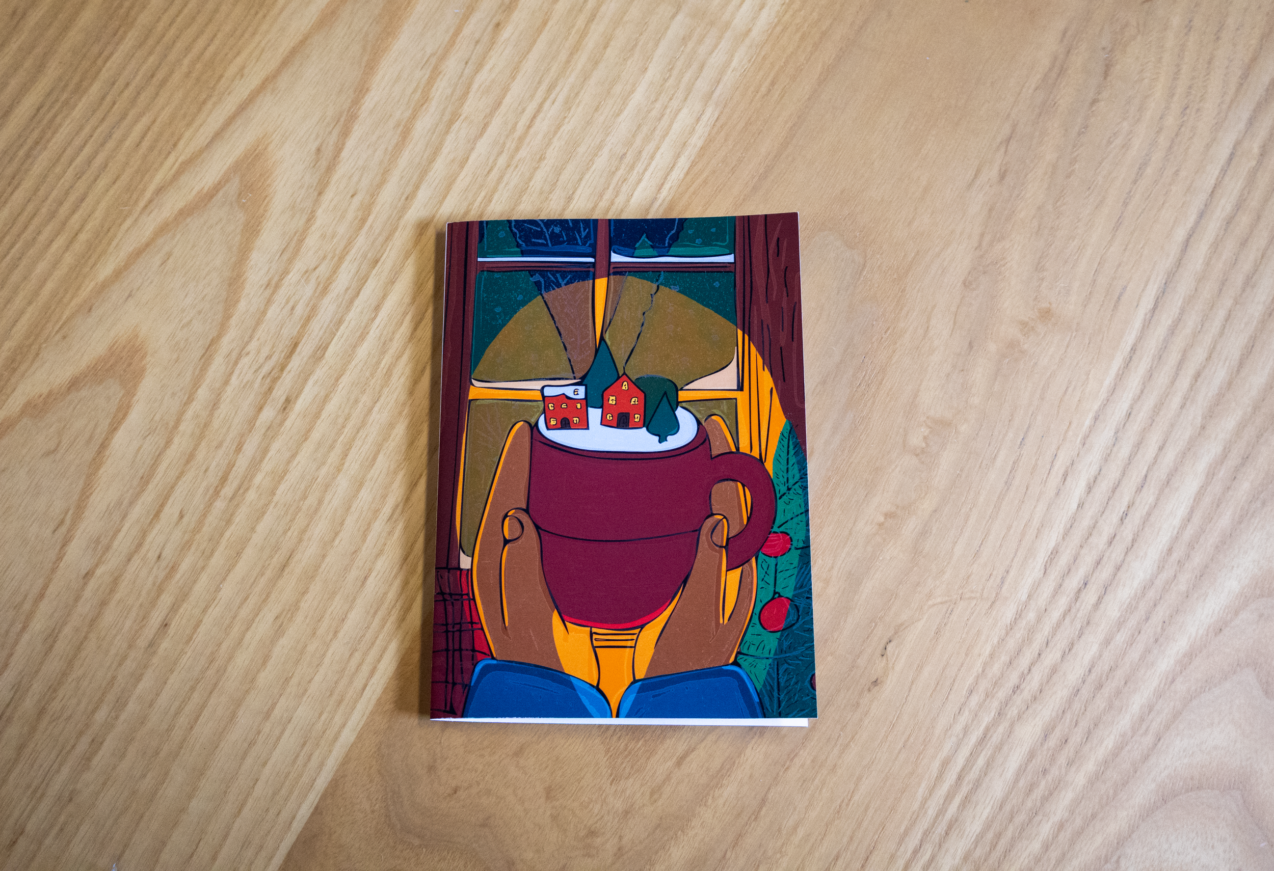 Carte illustrée posée sur une table en bois, représentant deux mains tenant une grande tasse chaude devant une fenêtre. À l’intérieur de la tasse, un petit village enneigé avec maisons et sapins.