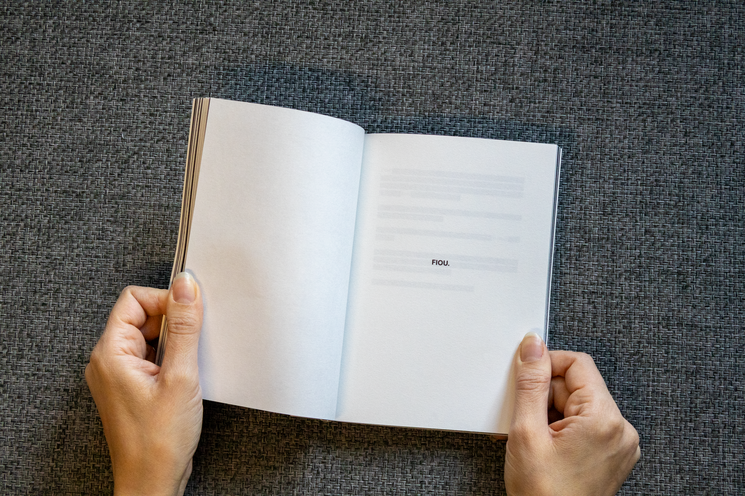 Une personne tient un livre ouvert avec une page blanche et une autre avec le mot 'FIOU' écrit dessus, posé sur une surface en tissu gris.