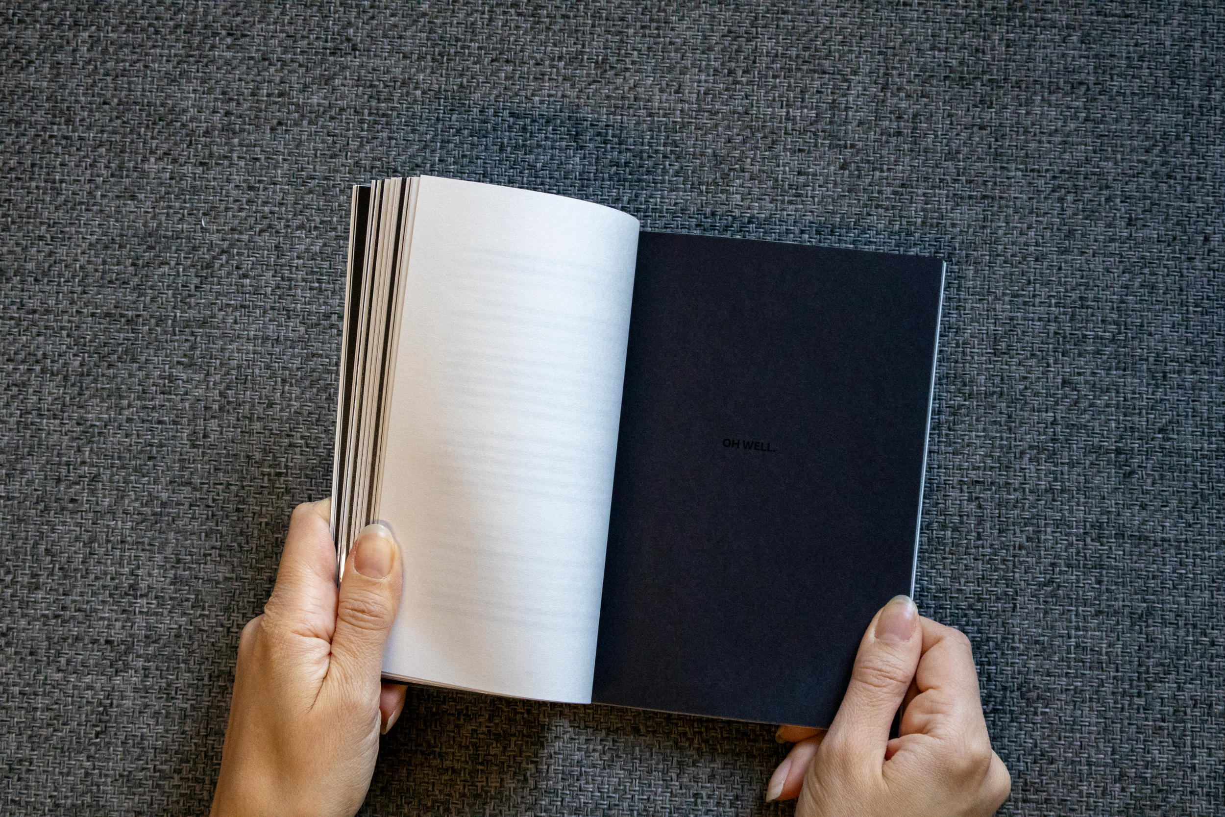 Une personne tient un livre ouvert avec des pages blanches d'un côté et une page noire avec le texte 'OH WELL.' au centre.