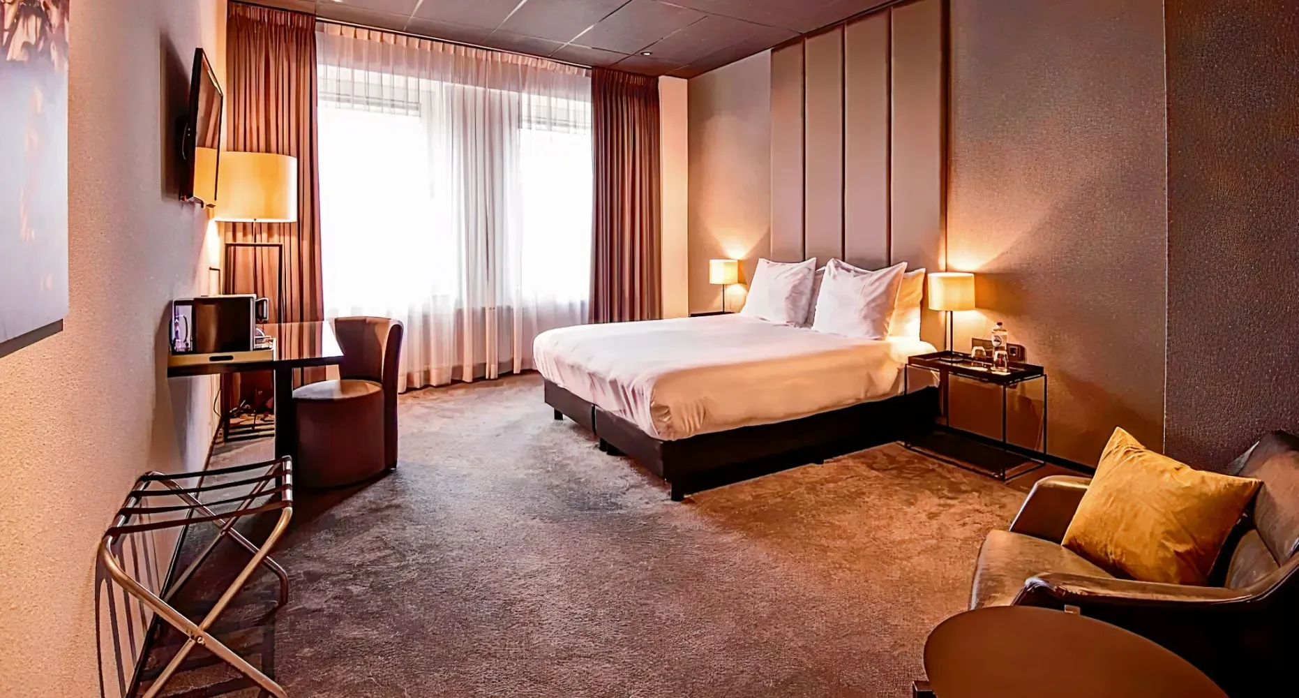 Een hotelkamer met een groot bed, beddengoed, nachtkastjes met lampen, een venster met gordijnen, een zithoek met stoel en een tafel, en een tv aan de muur.