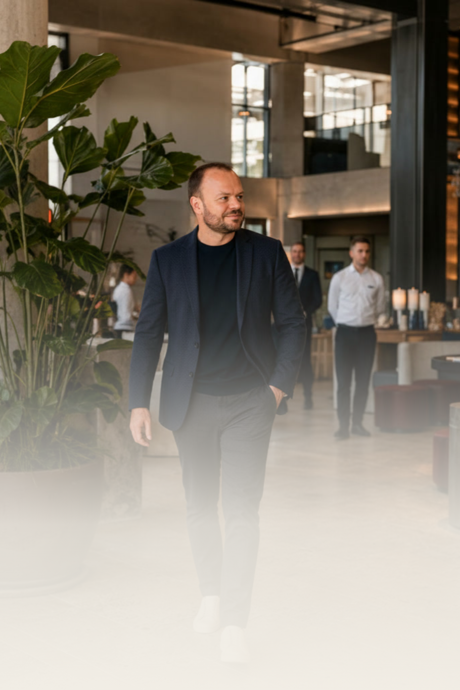 Man in zakelijke outfit wandelt door een interieur met planten en een bar, met andere mensen op de achtergrond.