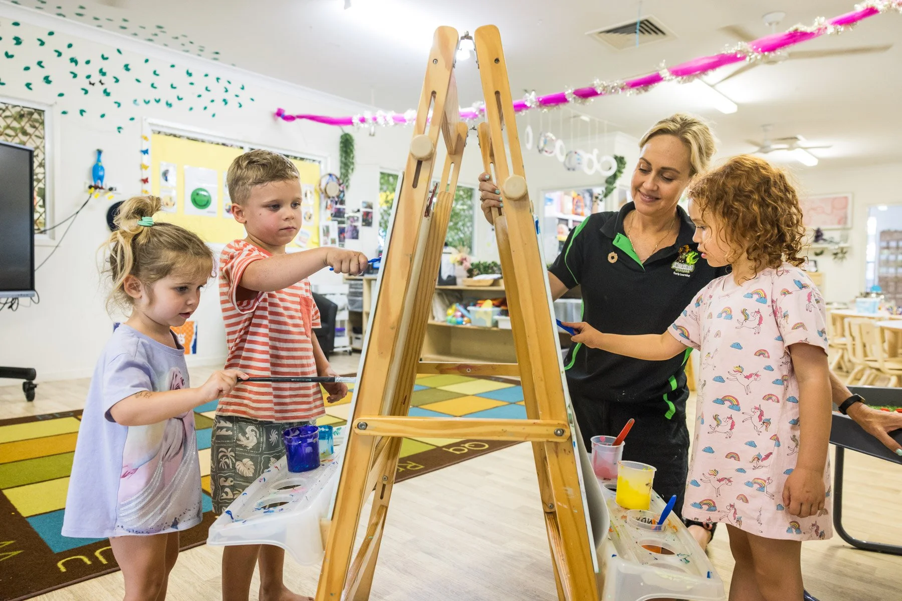 A young boy and an adult woman painting on a large canvas together in an art classroom.