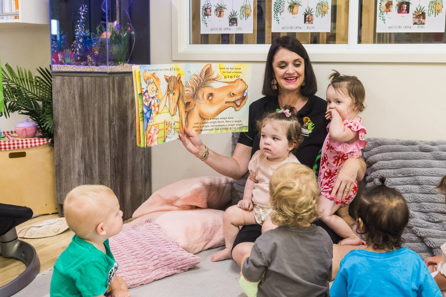 A woman reading a colorful children's book about farm animals to a group of young children in a cozy indoor setting.