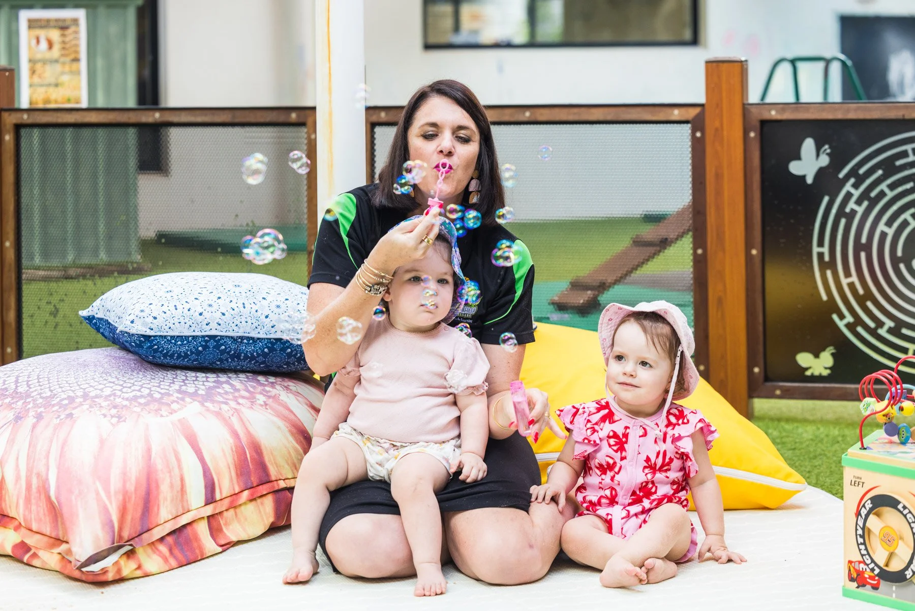 Woman and two young children sitting on the floor, with one girl blowing bubbles. The woman is holding a bubble wand, and the children are seated near pillows, with one girl wearing a sun hat and the other sitting on the woman's lap. There are toys and a bubble solution container beside them.