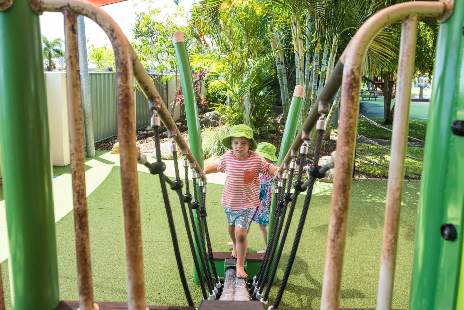 Children playing on a rope bridge at a playground with greenery and tropical plants in the background.