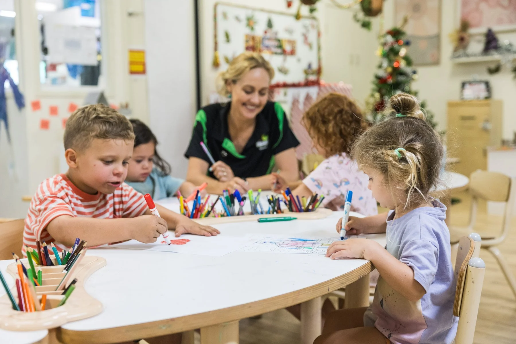 A group of young children and a female teacher sit around a table drawing and coloring with markers in a classroom decorated for Christmas, including a Christmas tree and holiday decorations.