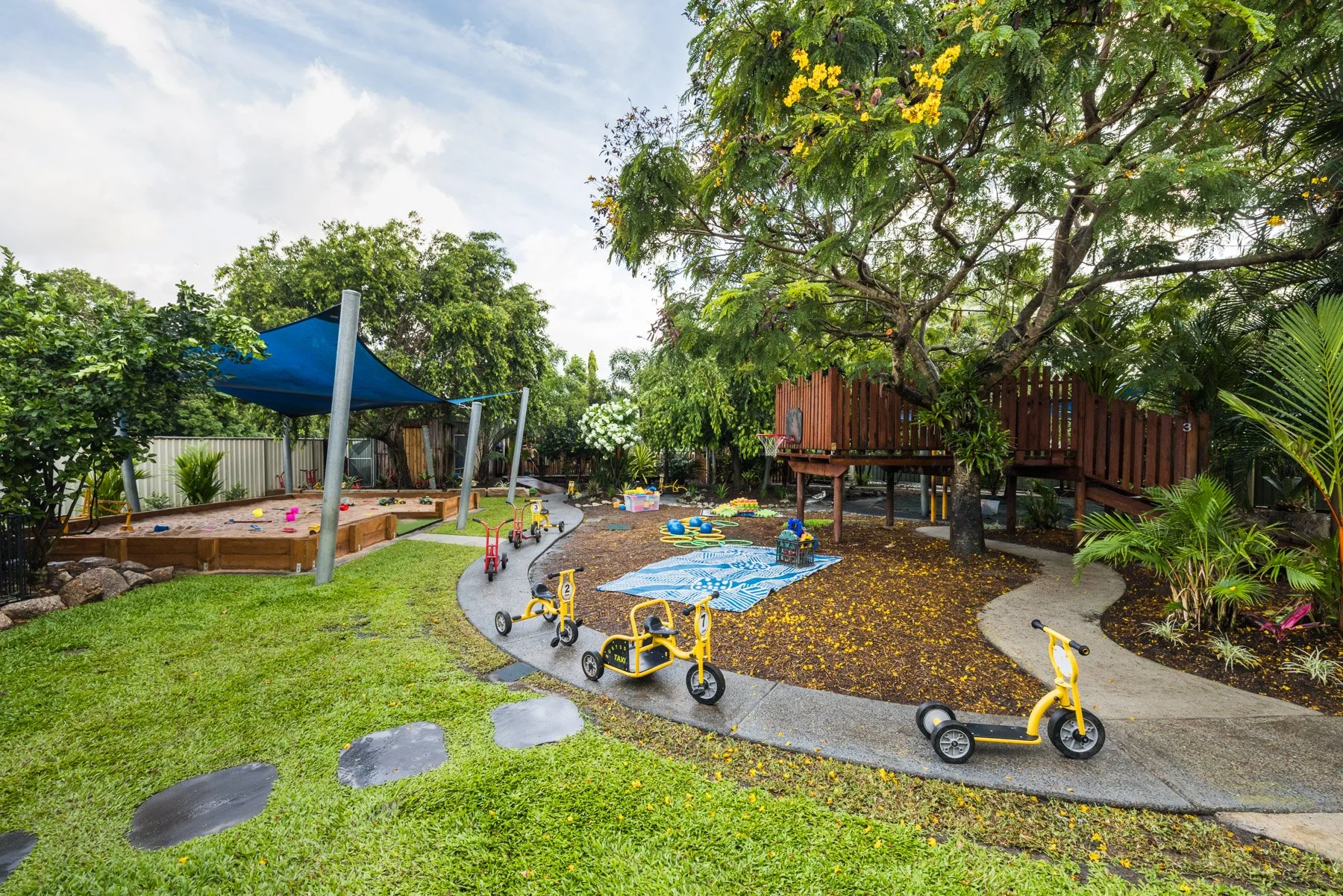 Children's outdoor play area with sandbox filled with toys, small slide, rooftop shade sails, hanging flower pots, and bordered garden with plants.