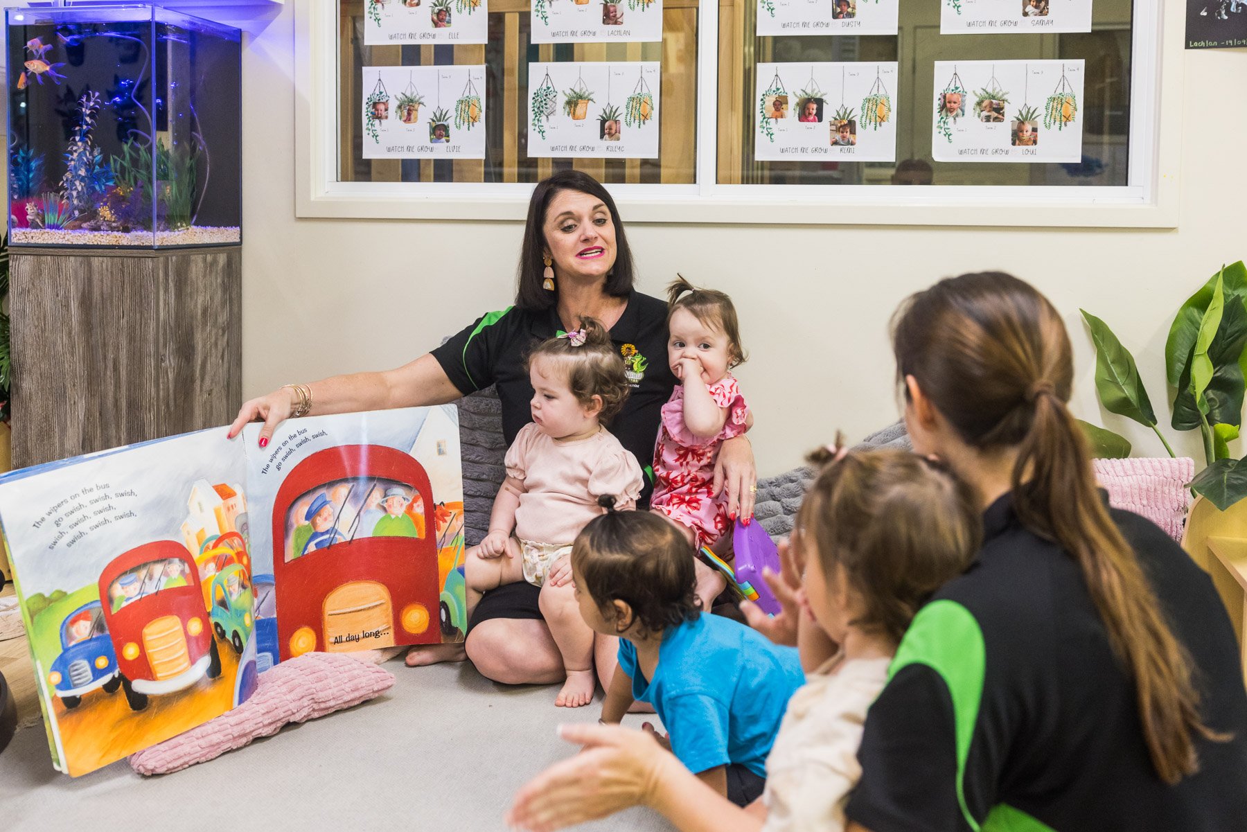 A woman reading a children's book to a group of young kids in a classroom or daycare setting. The woman is sitting on the floor with the kids, and the book shows illustrations of cars. The children are sitting on the floor, some with attentive or curious expressions.
