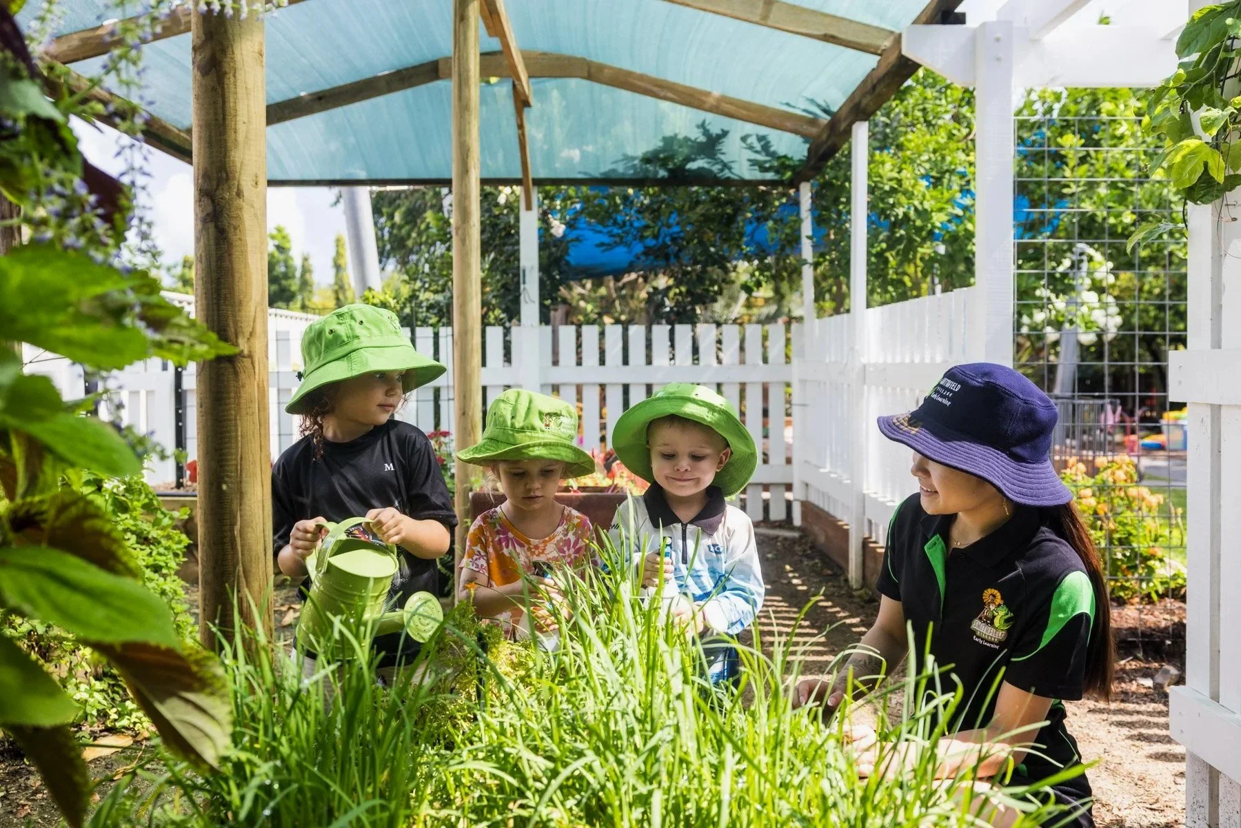 Children and a woman gardening in a small outdoor garden with a white picket fence, greenery, and sunlight.