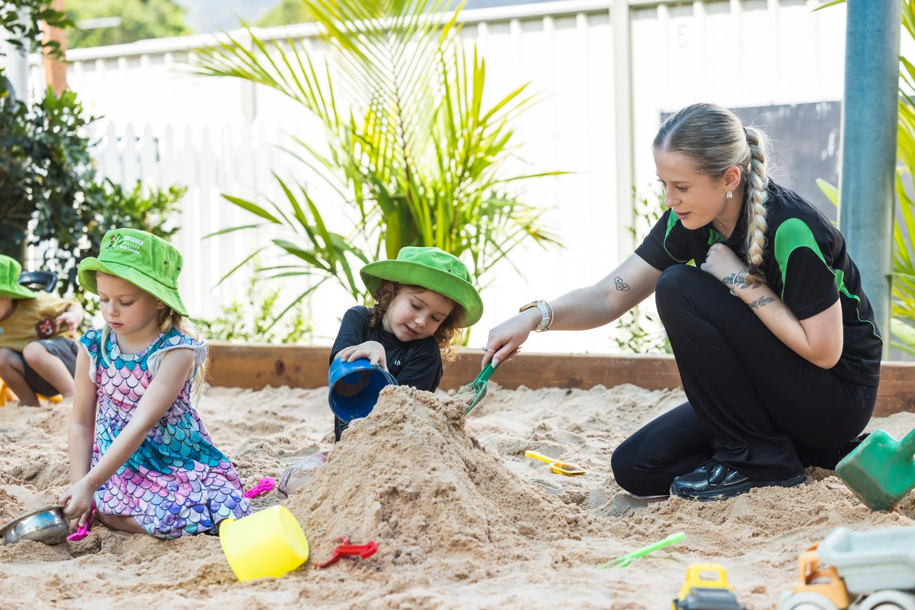 Children and an adult woman play in a sandbox with sand toys outdoors, with green plants and a white fence in the background.