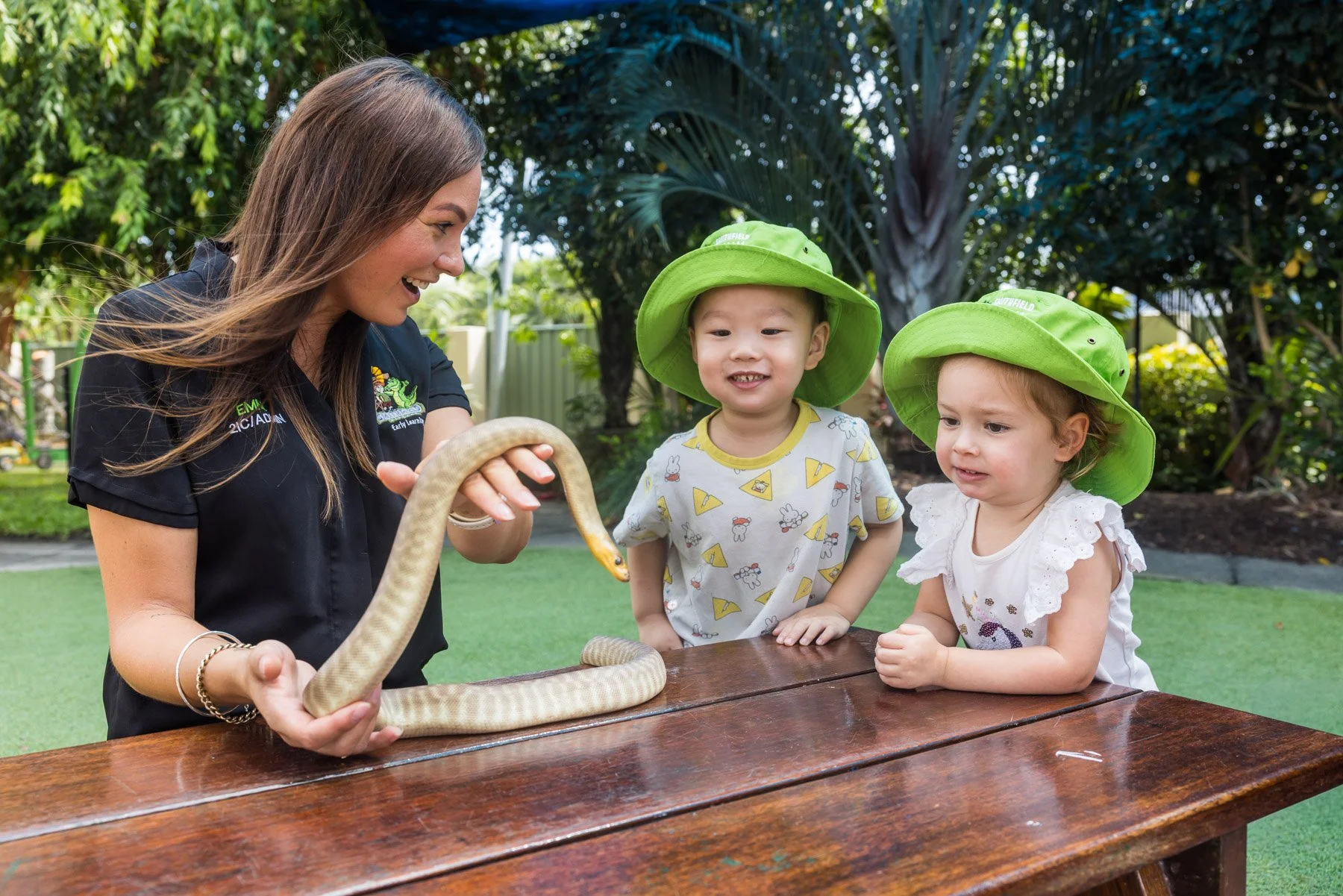 An adult woman holding a snake in front of two young children wearing green hats, outdoors in a garden setting.