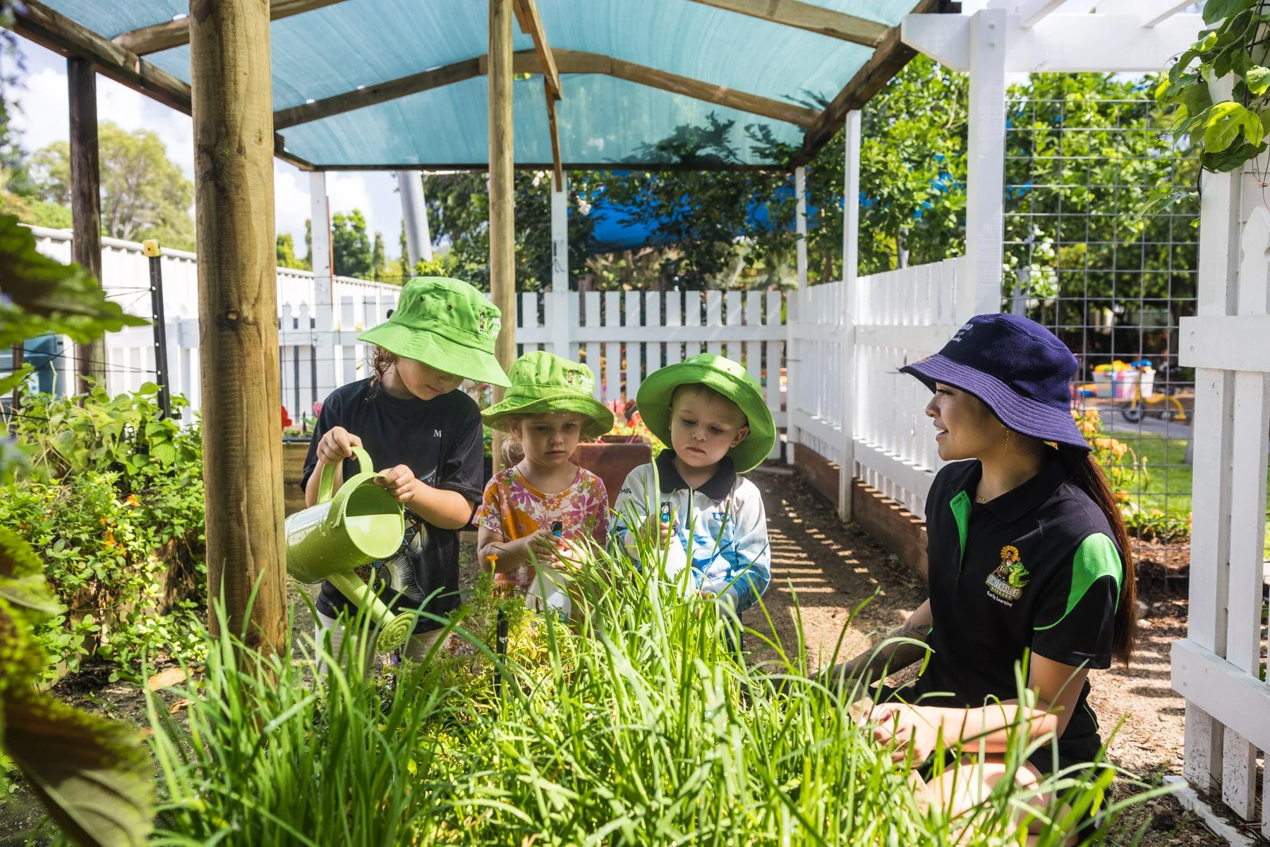 A woman and four children wearing hats, watering plants in a garden with white picket fence.