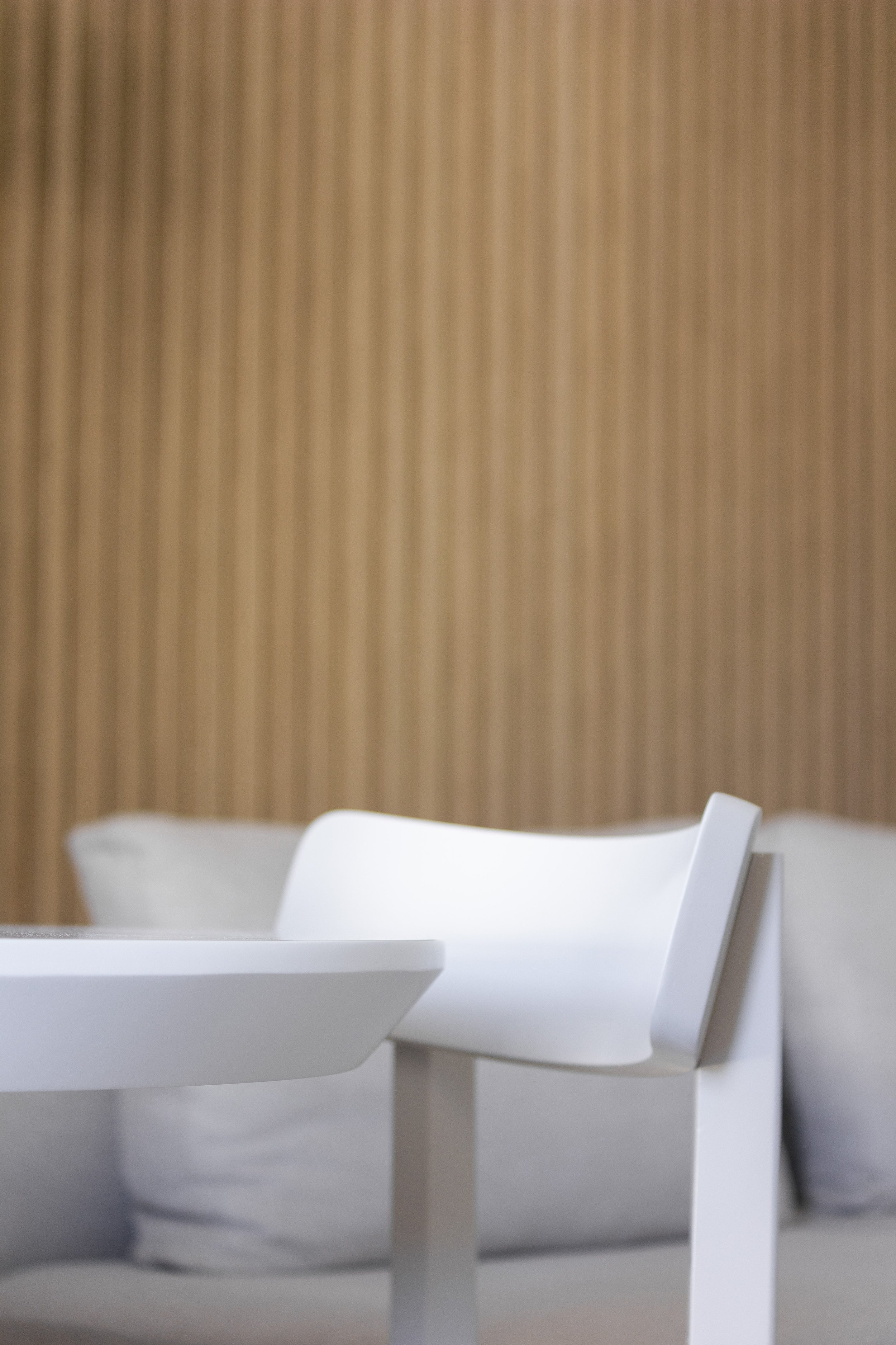 Cadeira branca ao lado de uma mesa branca, com almofadas cinzas ao fundo, parede de madeira listrada clara.