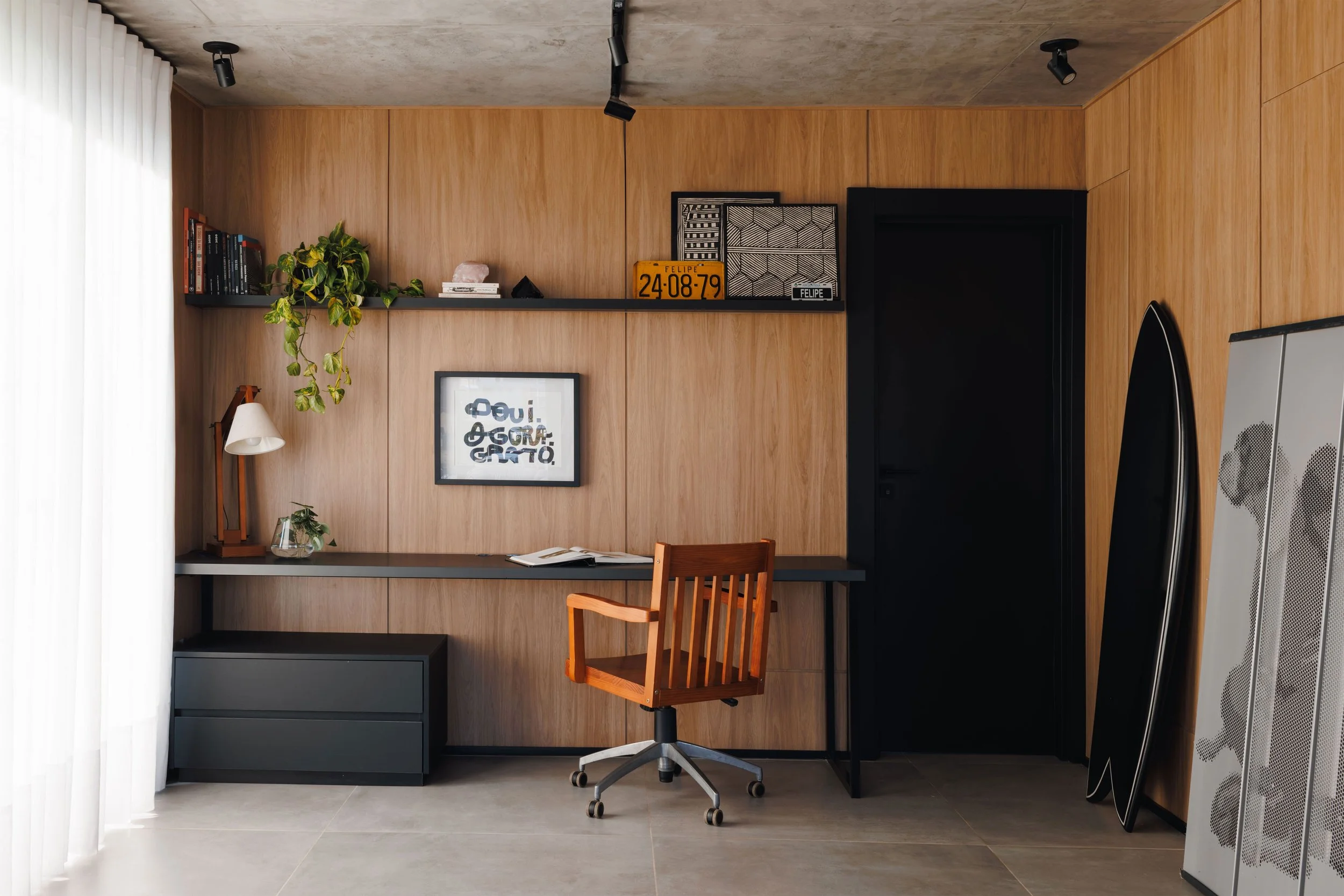 Escritório com painel de madeira na parede, mesa de estudos com livros e uma cadeira. Há uma luminária de mesa, uma planta, uma janela com cortinas brancas, prateleiras com livros, objetos decorativos, um quadro com a frase 'Aqui é o seu espaço feito