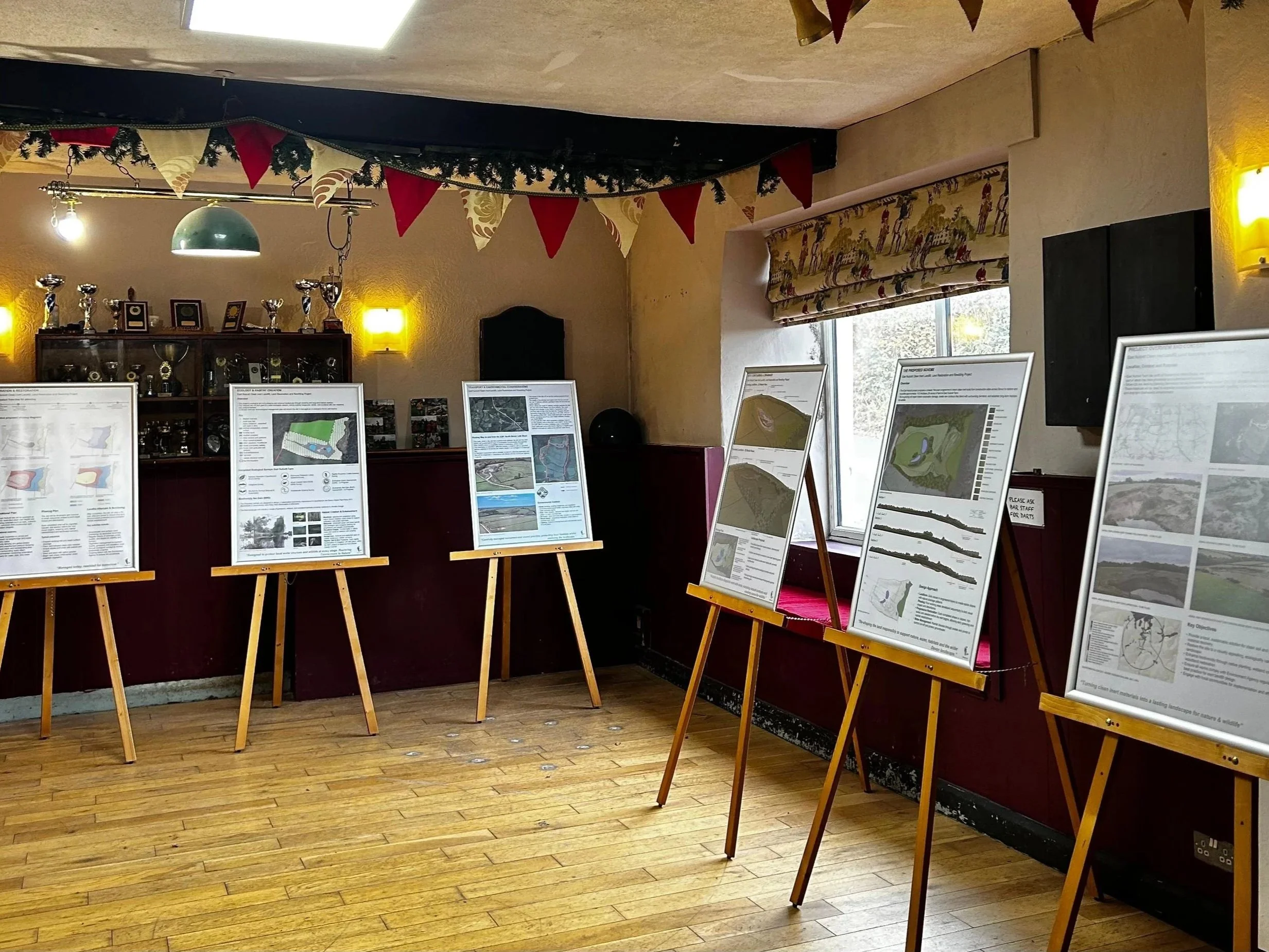 An indoor exhibition space with five display boards on easels showing informational posters about landscape and land use, a red and green bunting hanging from the ceiling, trophy cups and framed photographs on a shelf behind a counter, and a window providing natural light.