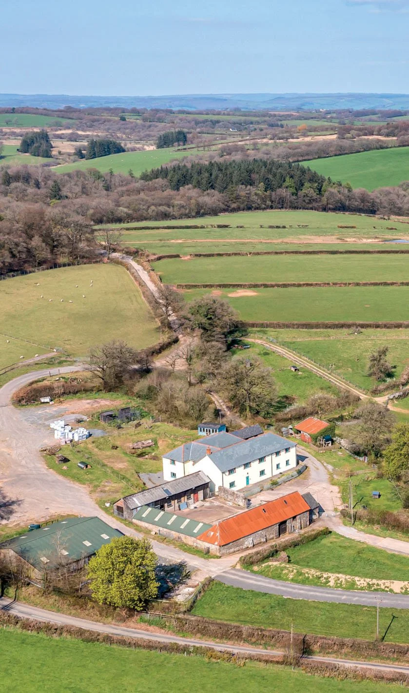 A rural landscape with farmland, fields, and scattered farm buildings, with rolling hills and trees in the background.