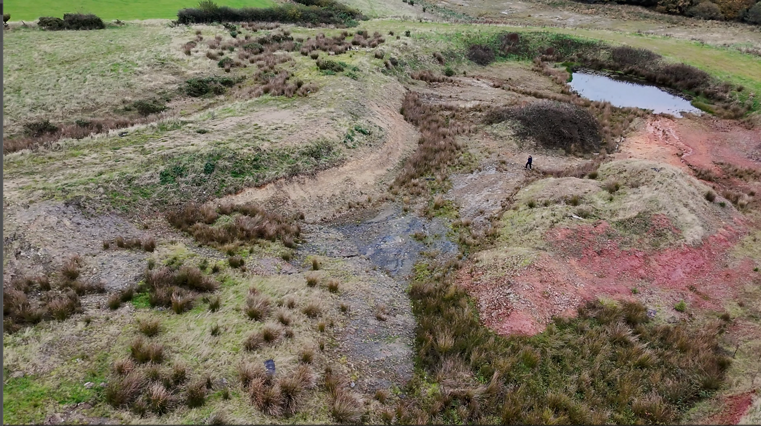 Aerial view of a landscape with patches of grass, shrubs, and small water bodies, with a person walking near a stream.
