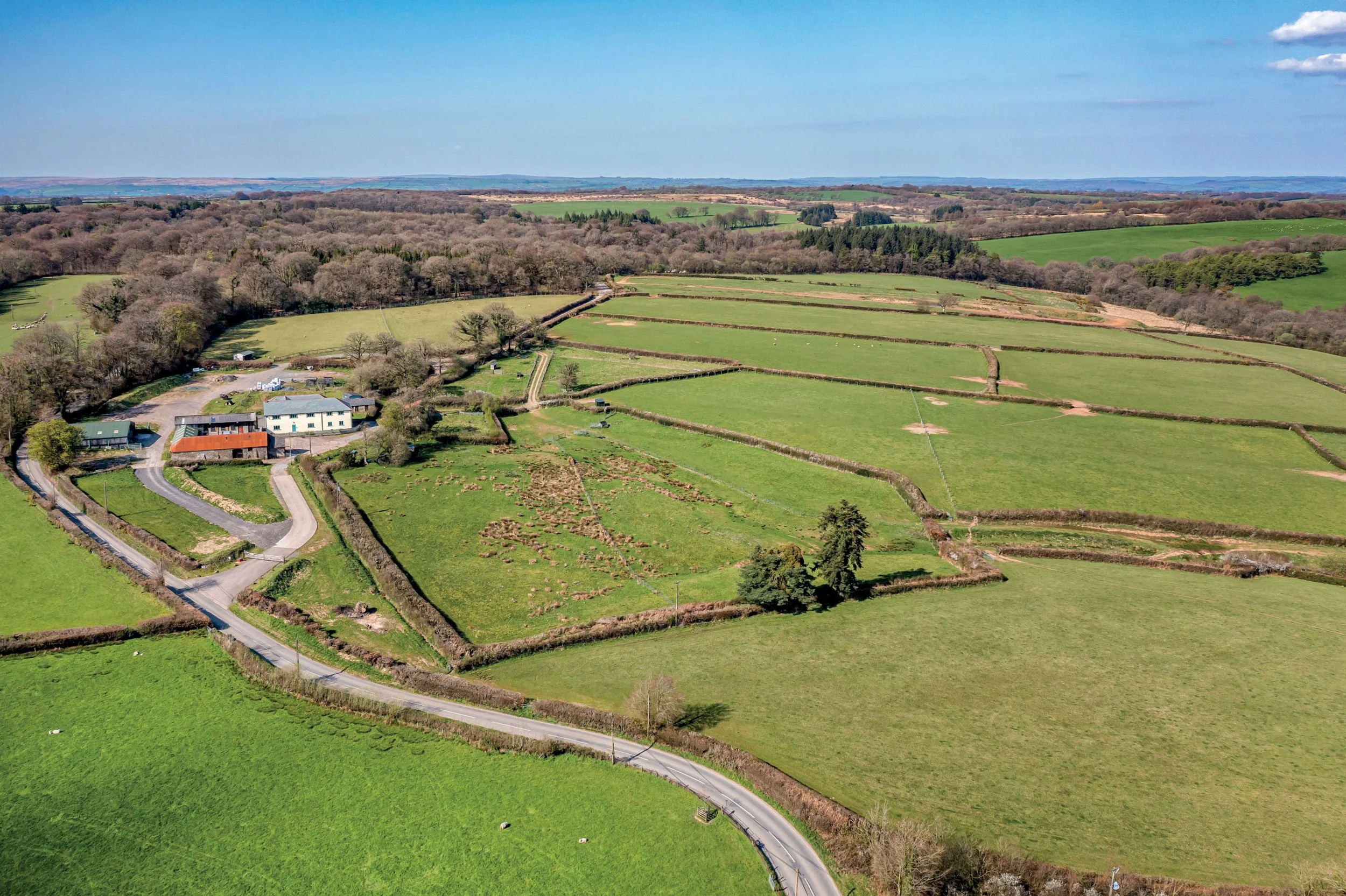 Aerial view of a green rural landscape with fields separated by stone walls, a few houses near the bottom left, and a winding road.