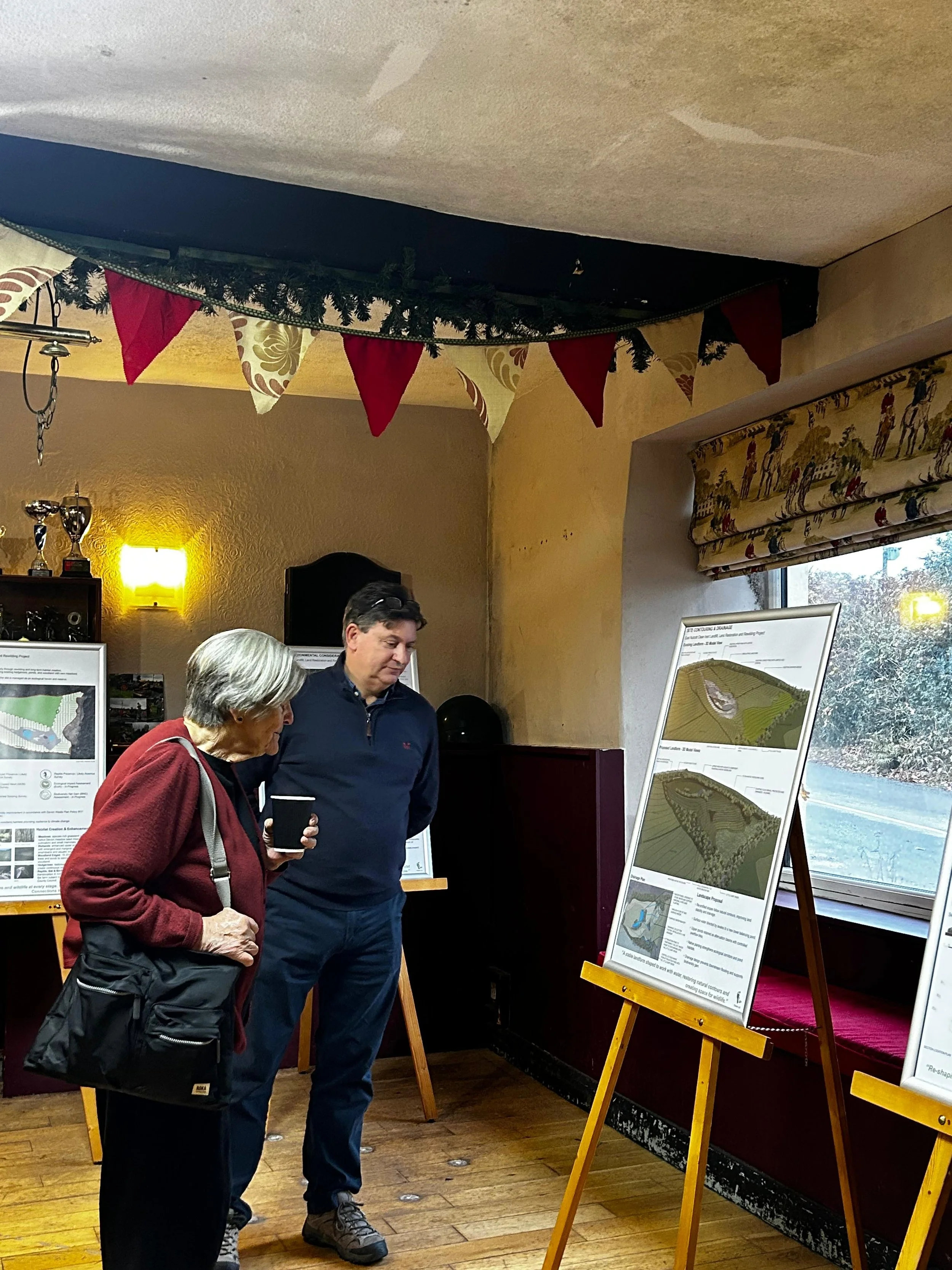 Two people examining detailed plans or maps on display boards inside a room decorated with bunting and trophies, with windows showing outdoor scenery.