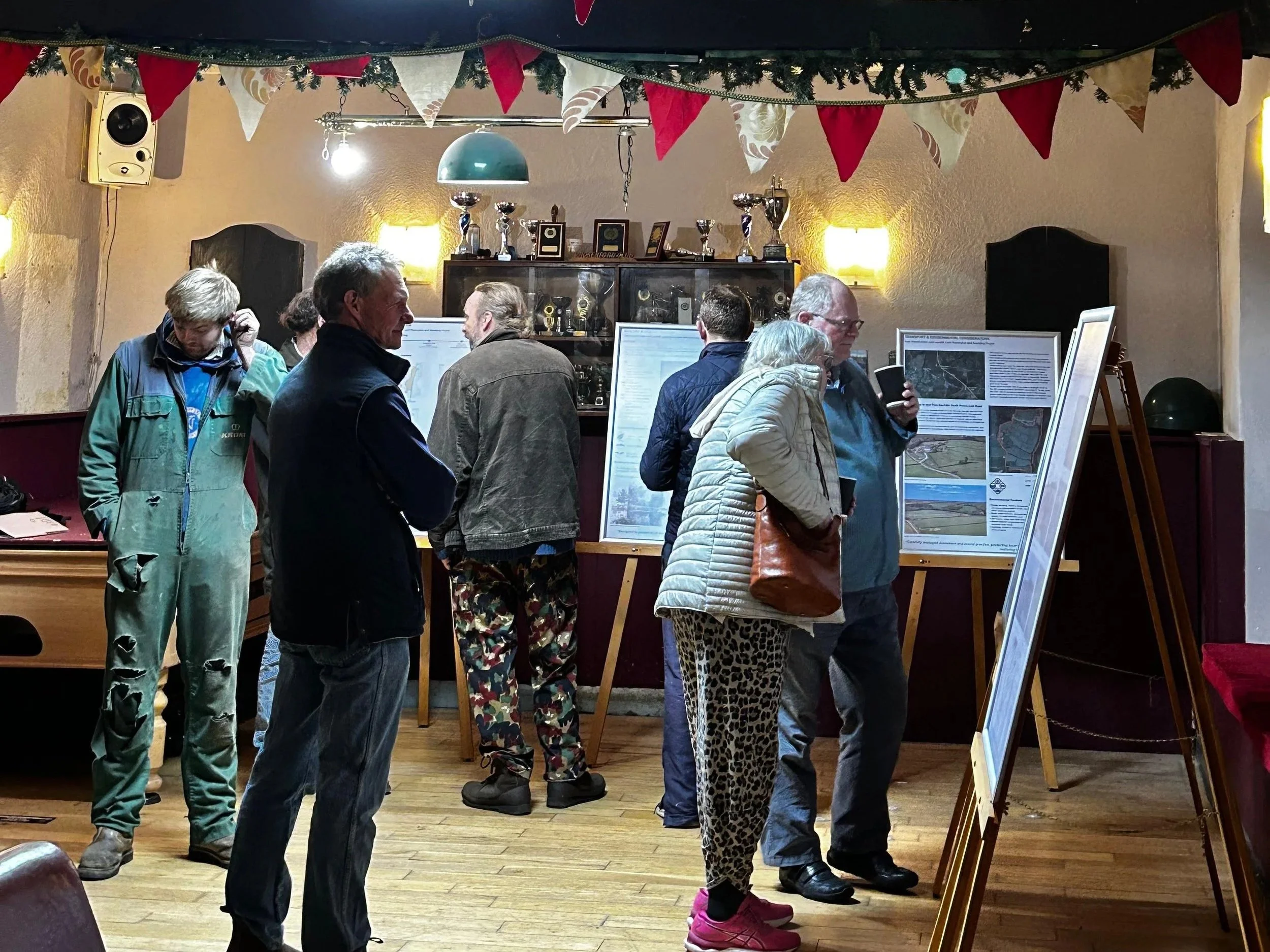 People viewing posters on display boards in a decorated indoor setting, with trophies on a cabinet in the background and bunting hanging from the ceiling.