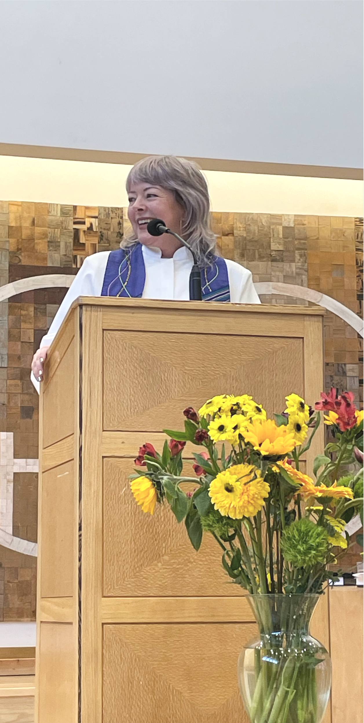 A woman with gray hair smiling and speaking at a podium inside a church or community hall, with a flower arrangement in the foreground.