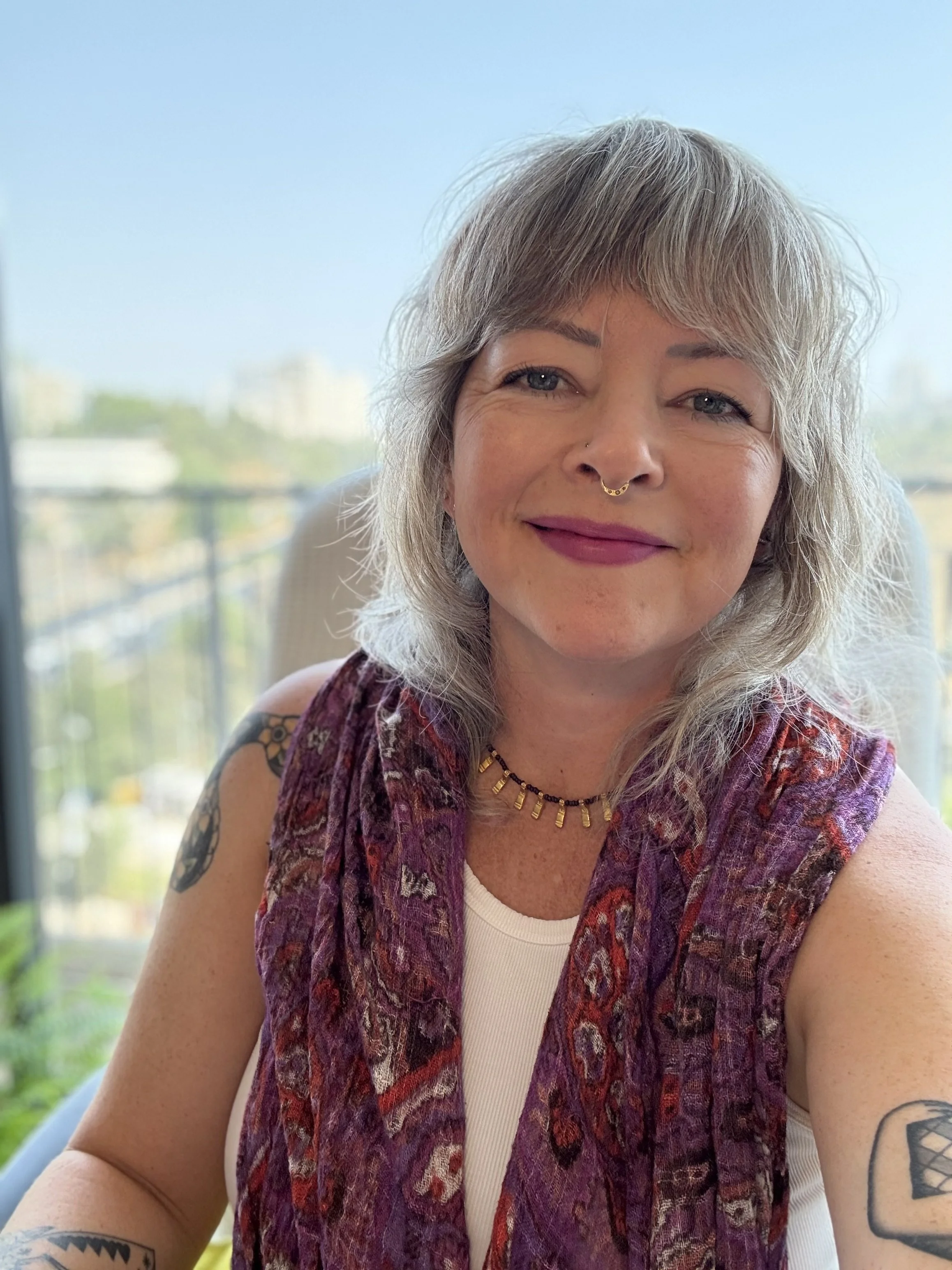 A woman with curly gray hair and blue eyes, wearing a septum piercing, a colorful scarf vest, a white tank top, a necklace, and tattoos on her arms, sitting in front of a window with a cityscape in the background.