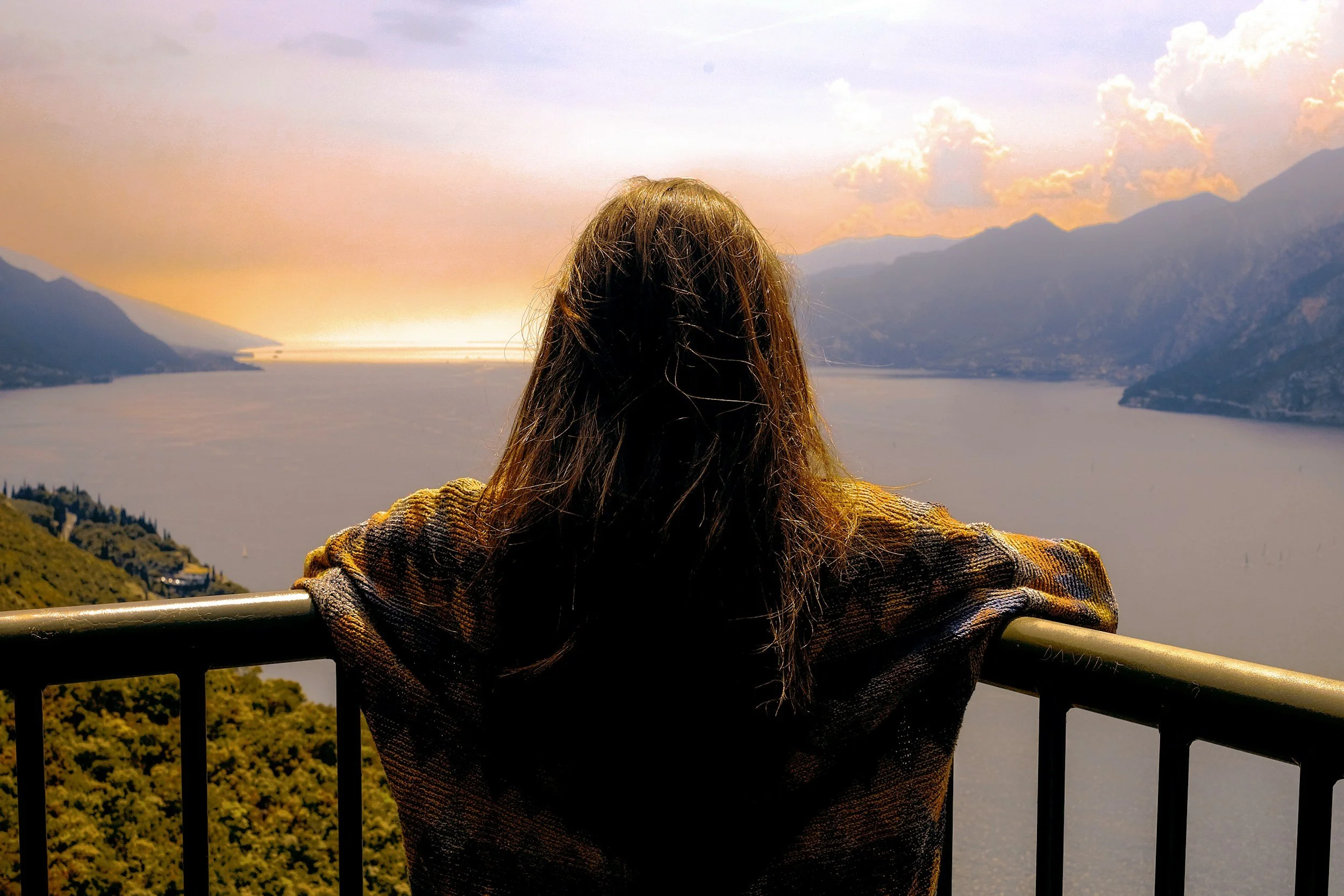 A person with long hair wearing a plaid jacket leaning on a railing and looking at a scenic lake view with mountains at sunset.