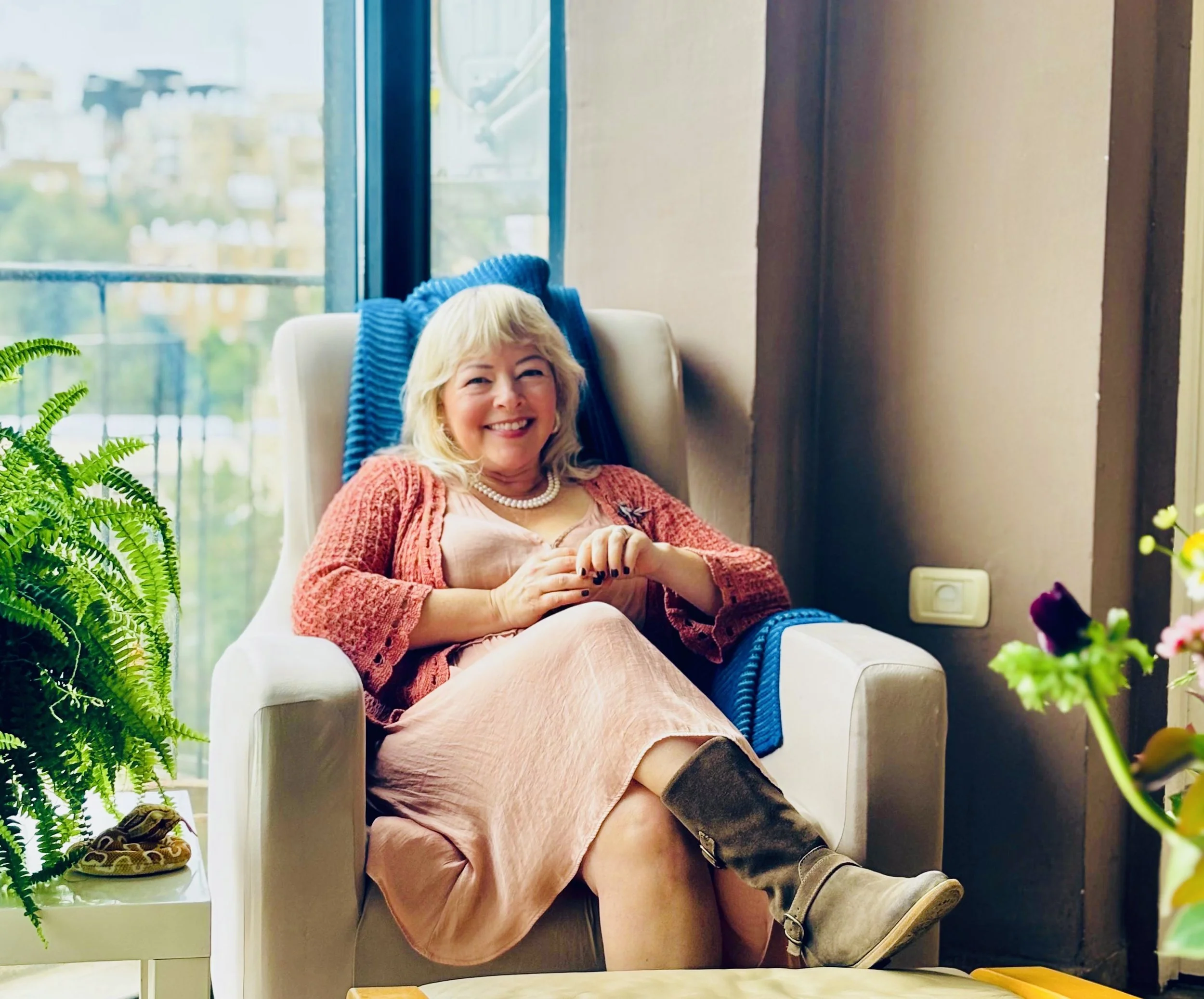 A woman with blonde hair smiling, sitting on a beige armchair by a large window. She is wearing a peach-colored dress, a pearl necklace, and a coral cardigan. There are green plants in the foreground and a snake on the table near her.