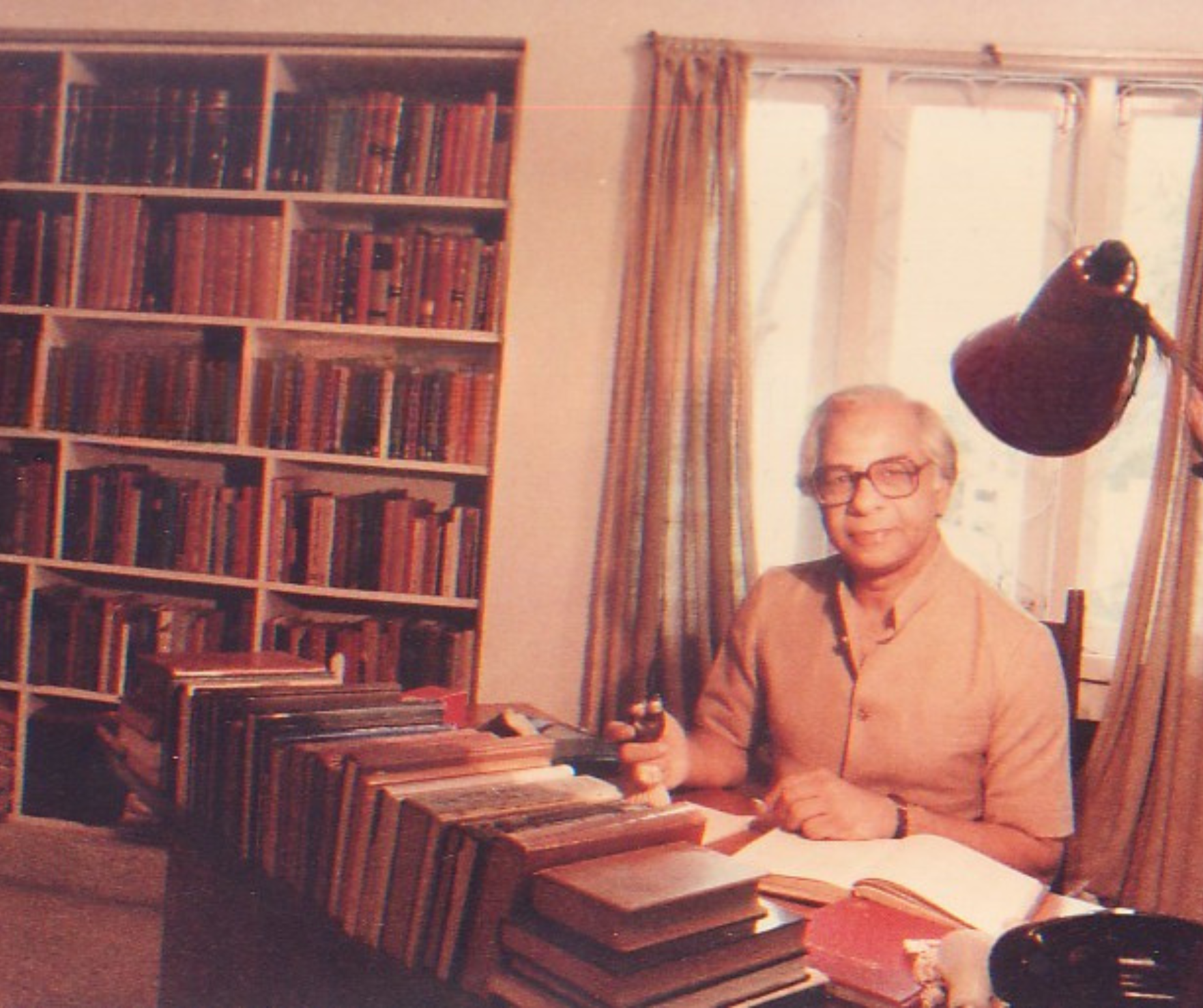 An elderly man with glasses and gray hair sitting at a desk in a room filled with books. He is holding a pipe and wearing a peach-colored shirt. Behind him is a large window with yellow curtains, and a reading lamp is attached to the window frame. There are many books stacked on the desk and arranged on a bookshelf in the background.