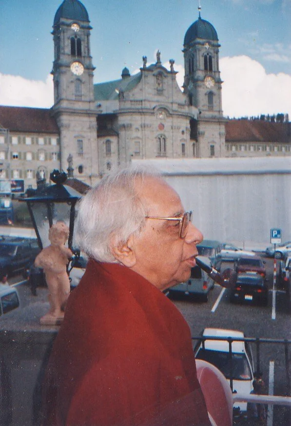 An elderly man with white hair and glasses smoking a pipe, standing outdoors with a historic church building and parked cars in the background.