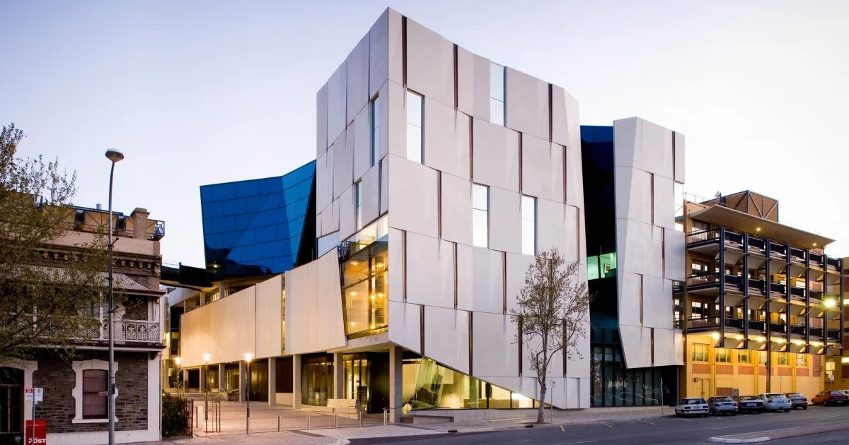 Modern multi-story building with an angular, abstract exterior design featuring large panels and windows, with some blue and white elements, located next to older brick buildings, shown during dusk or evening.