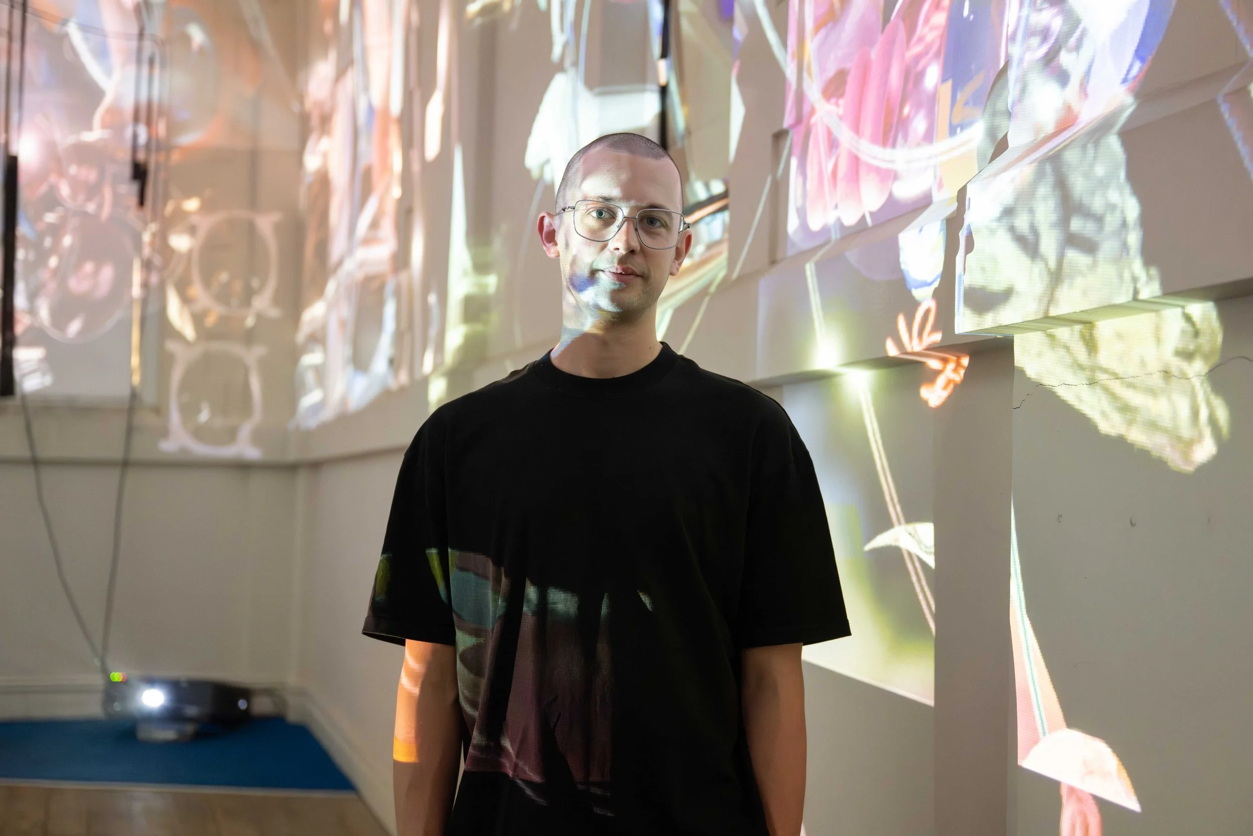 A person with glasses and a black t-shirt standing in front of a wall with digital artwork or projections of abstract and floral patterns.