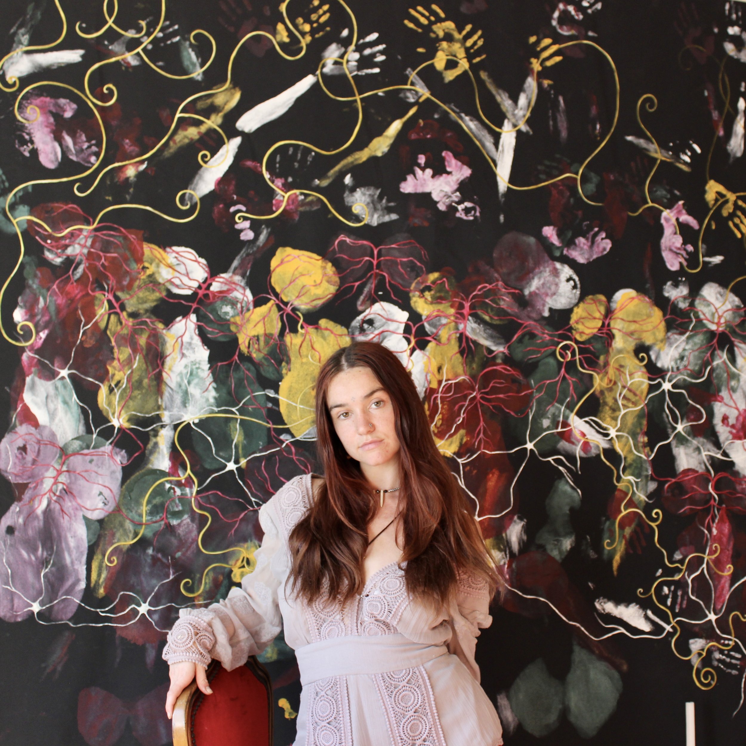 A young woman with long brown hair and a neutral expression, wearing a beige dress with lace details, sitting on a red chair in front of a colorful, abstract floral painting with black background and bright yellow, pink, and white accents.