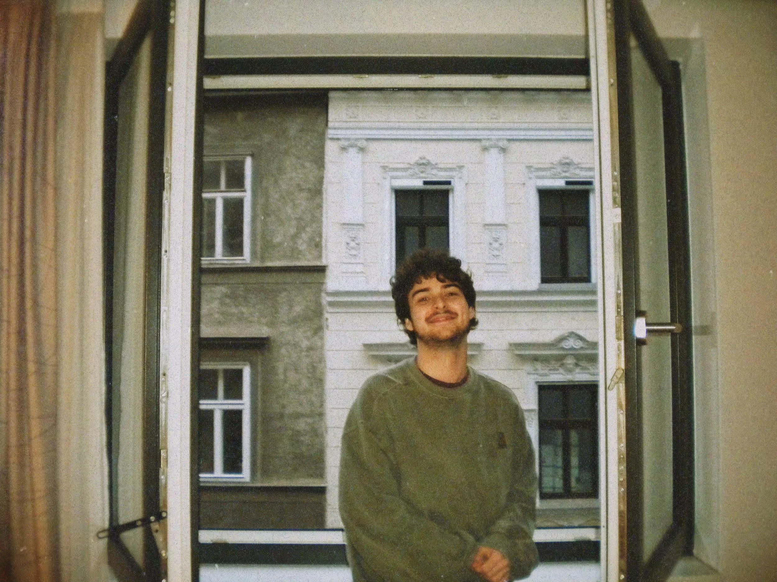 Ein junger Mann lächelt vor einem offenen Fenster, im Hintergrund ein altes, mehrstöckiges Gebäude mit weißen Verzierungen.