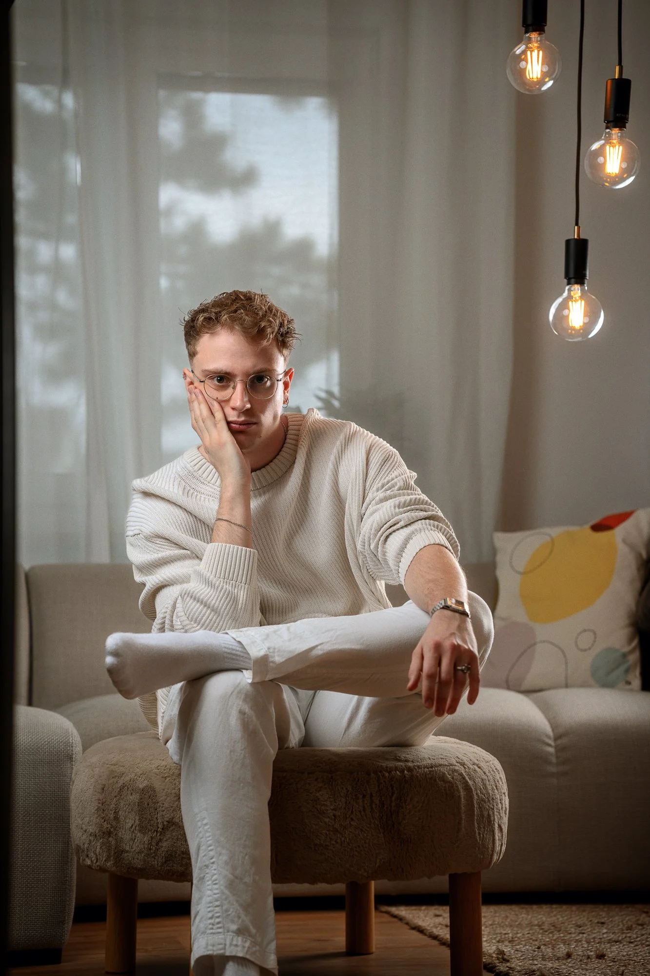 Ein Mann sitzt nachdenklich auf einem kleinen Hocker in einem Wohnzimmer, trägt eine cremefarbene Strickjacke und weiße Hosen, im Hintergrund sind ein Sofa, ein Kissen mit buntem Muster und eine Stehlampe mit Edison-Glühbirnen sichtbar.