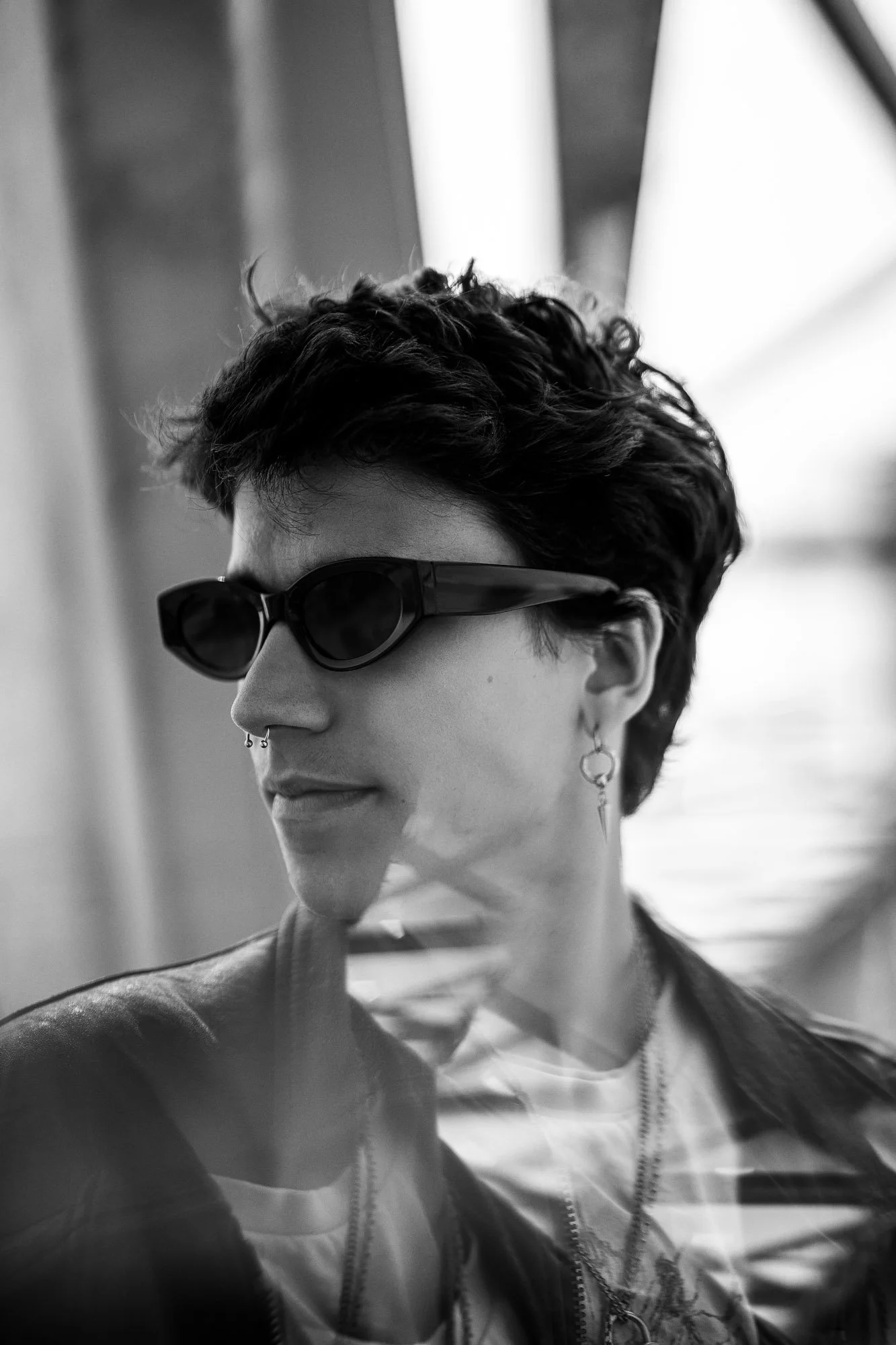 Schwarz-weiß-Foto einem jungen Mann mit kurzen, lockigen Haaren, Sonnenbrille, Piercings und Lederjacke, der nach links schaut, vor einem Fenster im Hintergrund.