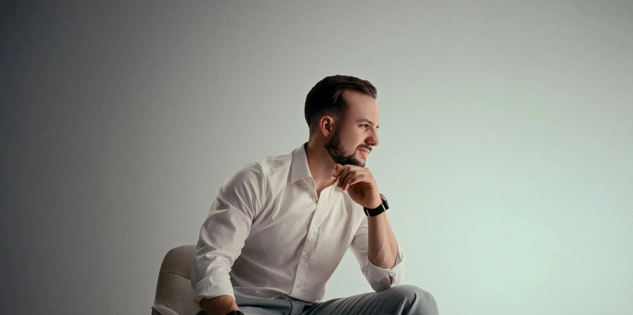 Ein Mann in einem weißen Hemd sitzt nachdenklich an einem Tisch, mit der Hand am Kinn und blickt nach rechts, vor einem hellen Wandhintergrund.