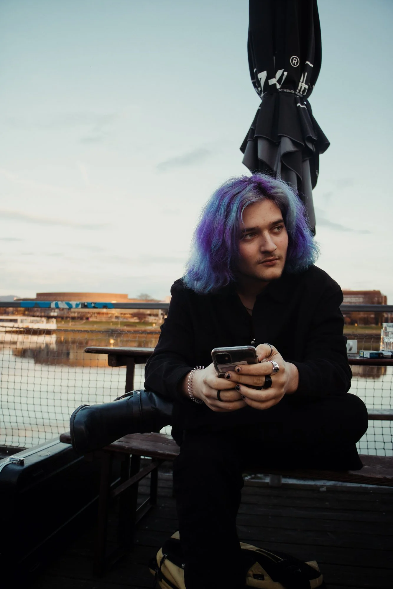 Junge Person mit lila- und blautönigem Haar sitzt an einem See bei Sonnenuntergang, hält ein Smartphone in den Händen und trägt schwarze Kleidung.