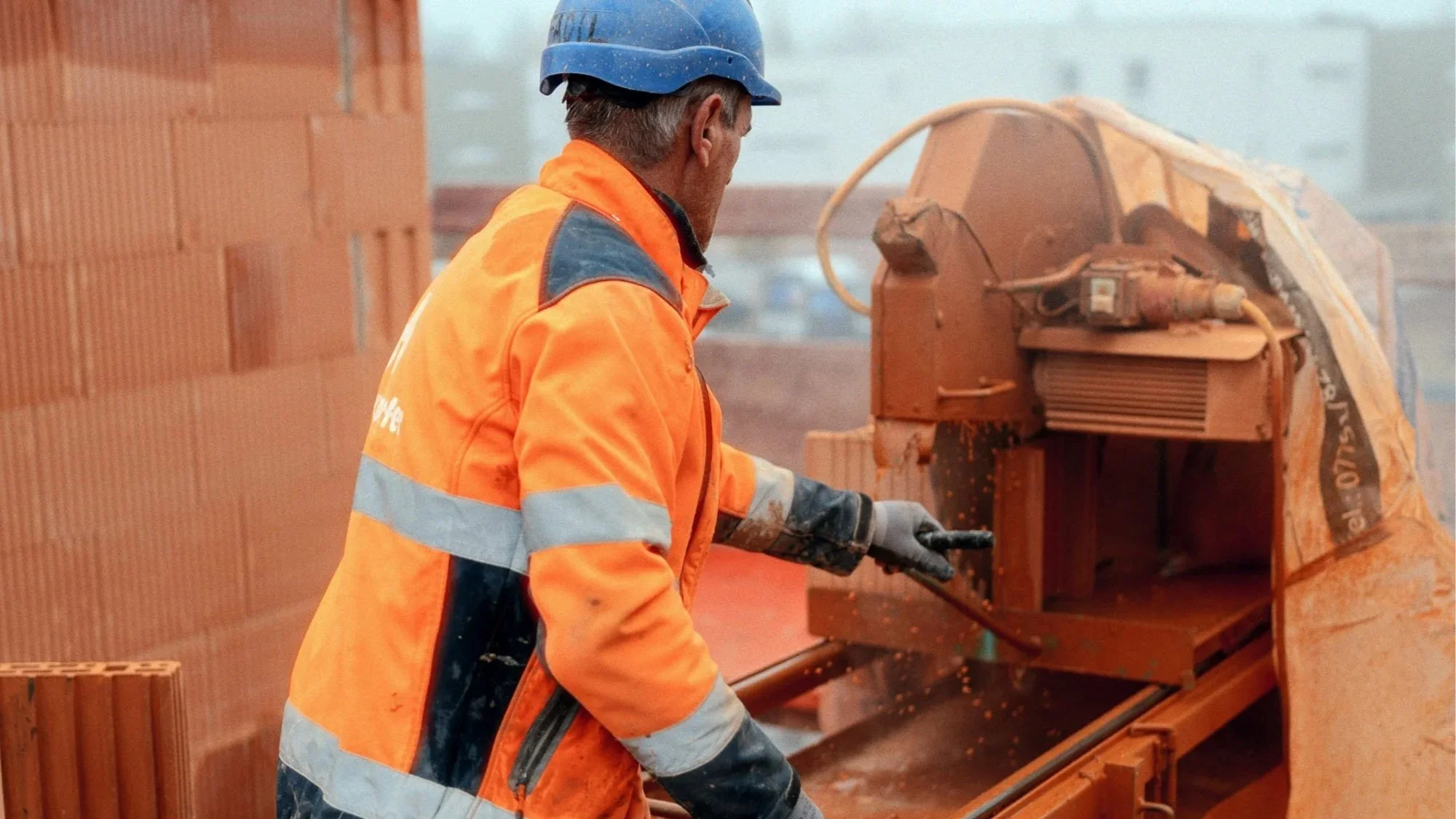 Bauarbeiter sammelt Ziegelsteine auf einer Baustelle mit einer technischen Maschine im Hintergrund.