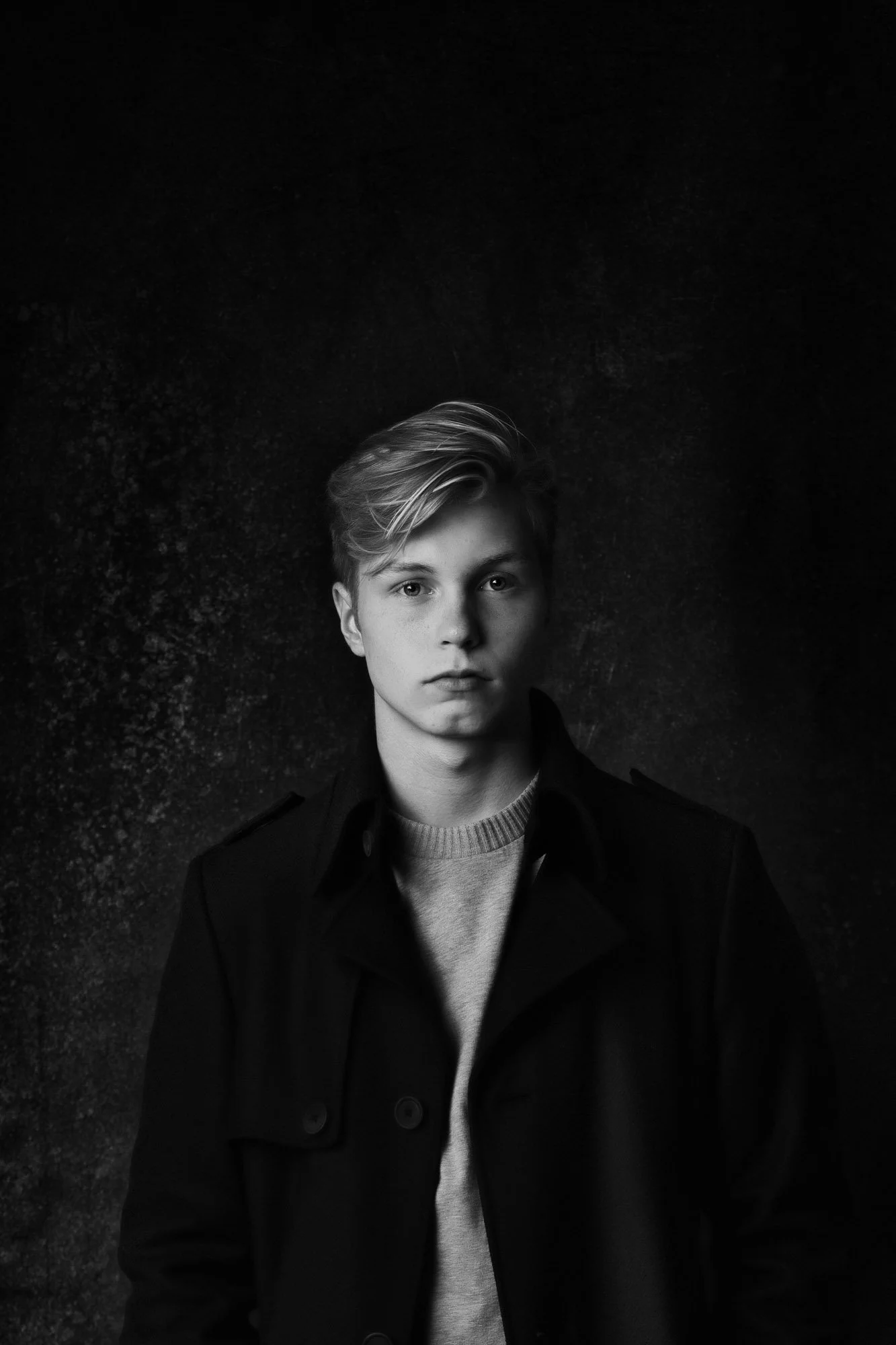 Schwarz-weiß-Porträt eines jungen Mannes, der in die Kamera schaut, mit kurzen, lockeren Haaren, trägt eine Jacke und einen Pullover, dunkler Hintergrund.