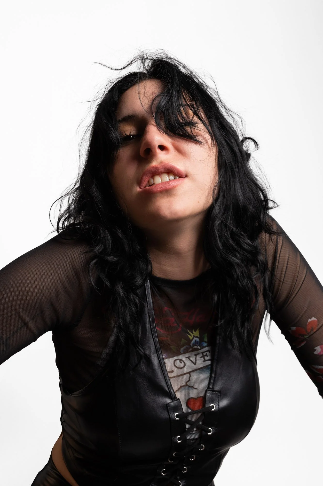 Junge Frau mit schwarzen, gewellten Haaren und dunklem Make-up, trägt ein schwarzes, durchsichtiges Oberteil und ein schwarzes Korsett, posiert vor weißem Hintergrund.