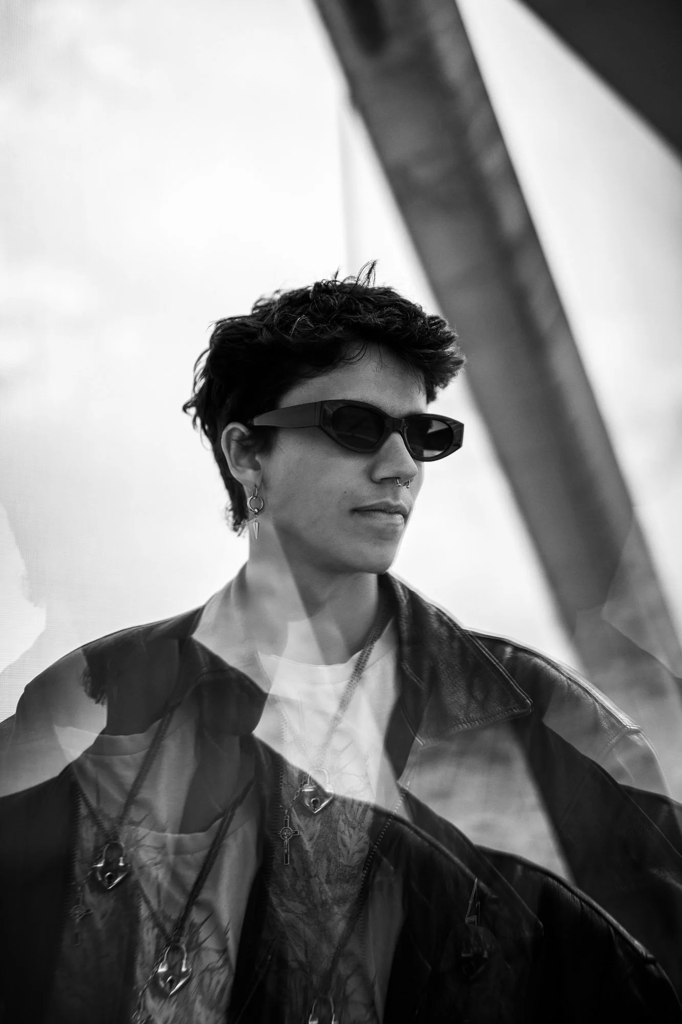 Schwarz-weiß-Foto eines jungen Mannes mit kurzen dunklen Haaren, Sonnenbrille, Lederjacke und mehreren Halsketten, mit reflektierender Glasscheibe vor ihm, die eine doppelte Darstellung seiner Person zeigt.