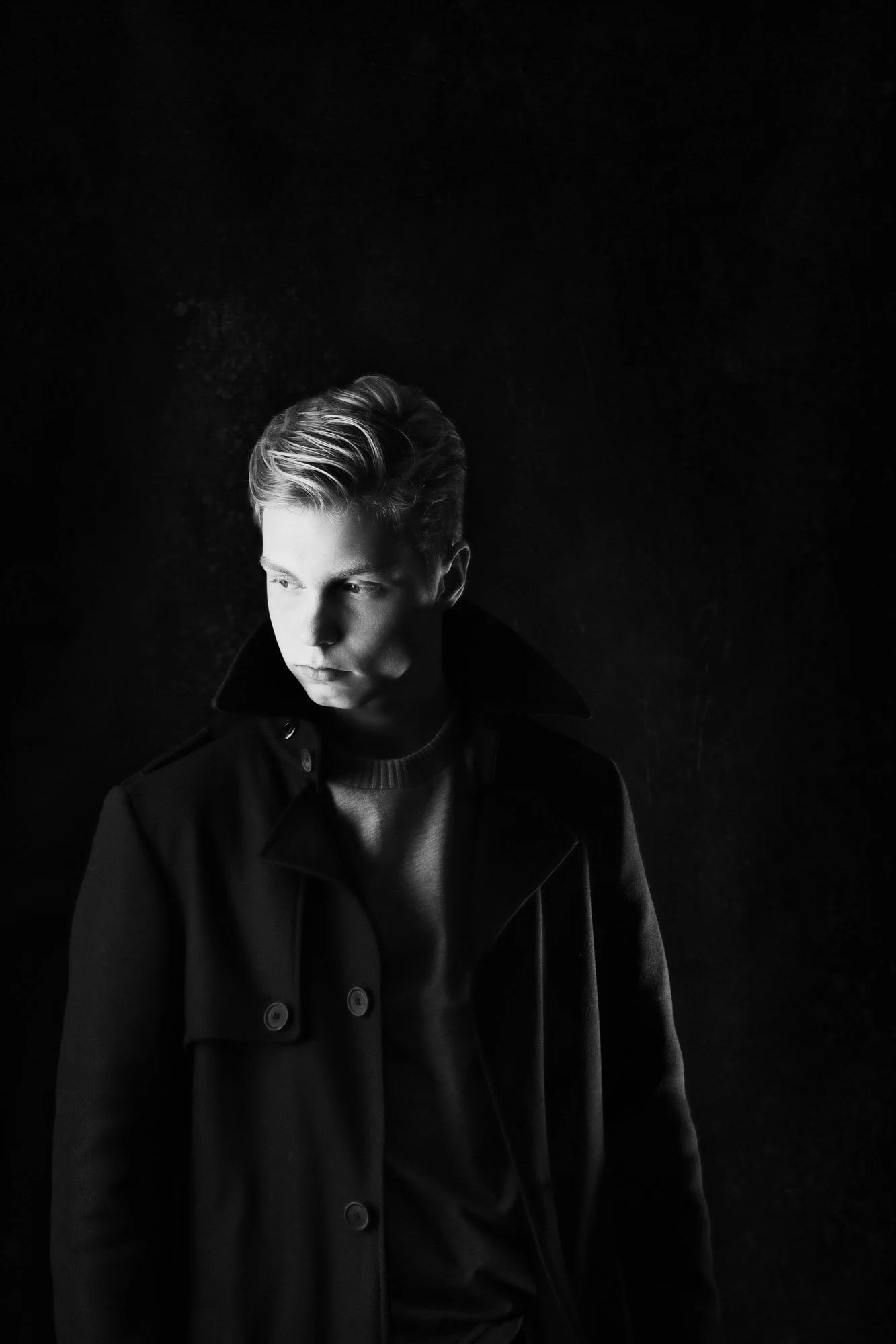 Schwarz-weiße Aufnahme eines jungen Mannes mit gepflegtem, geflämmtem Haar, der eine dunkle Jacke trägt und ins Licht schaut, mit dunklem Hintergrund.