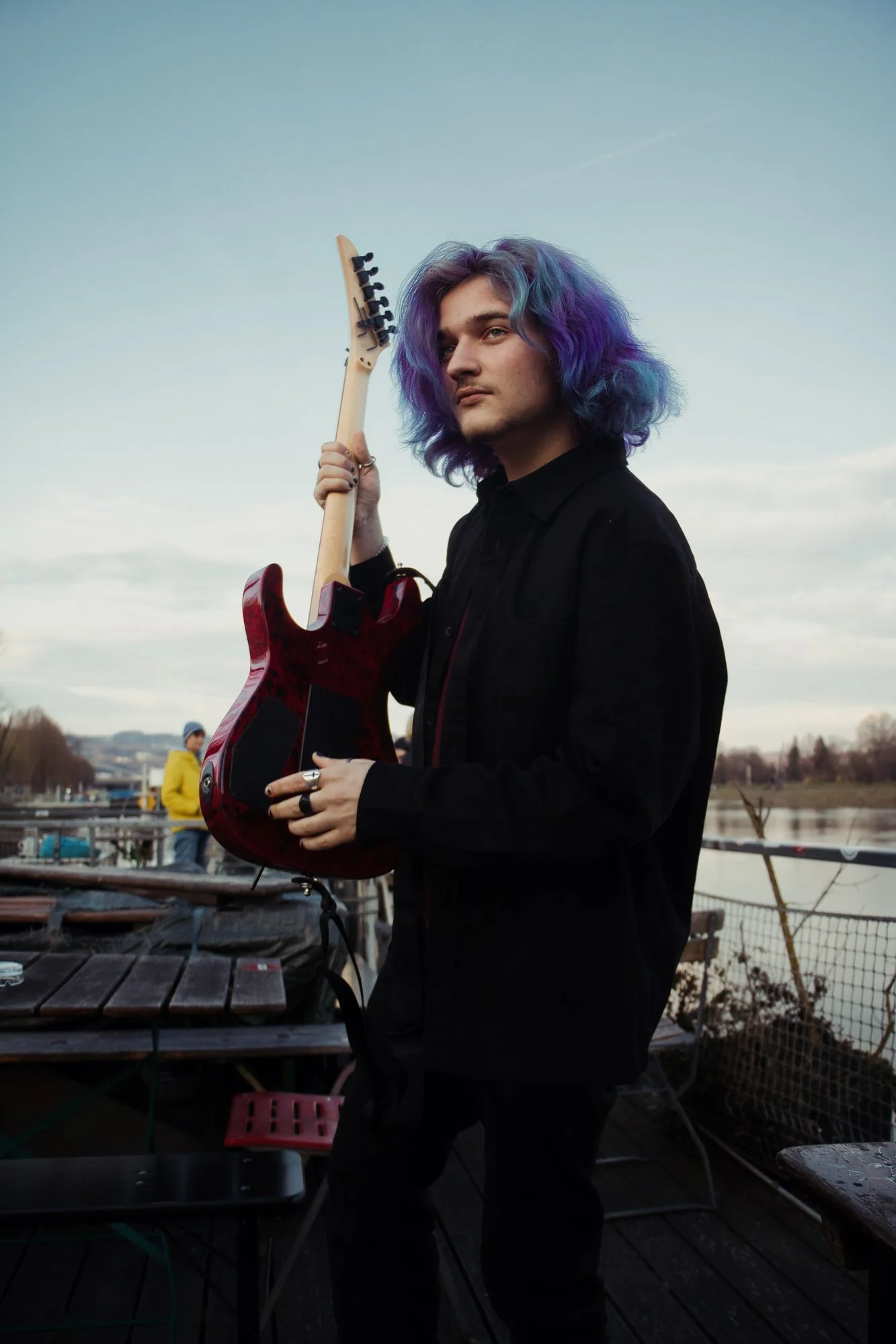 Porträt eines jungen Mannes mit lilafarbenem, lockigem Haar, der eine elektrische Gitarre hält, im Freien an einem Wasserufer bei leicht bewölktem Himmel.